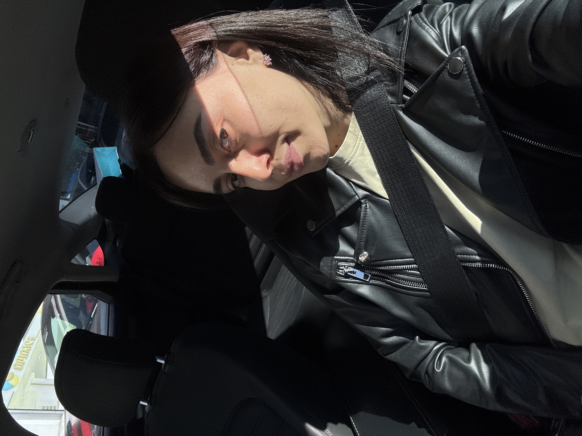 A young woman sits inside a car, presumably in Tbilisi, Georgia, based on the visible storefronts outside the car window. She wears a black leather jacket over a light-colored top and has her seatbelt fastened. The woman has dark hair and is wearing pink flower-shaped earrings. The interior of the car is primarily black, with the headrest and seat visible behind her. The sunlight streams through the car window, illuminating the woman's face. Outside the car, a sign in Georgian is visible, and storefronts suggest a commercial area.