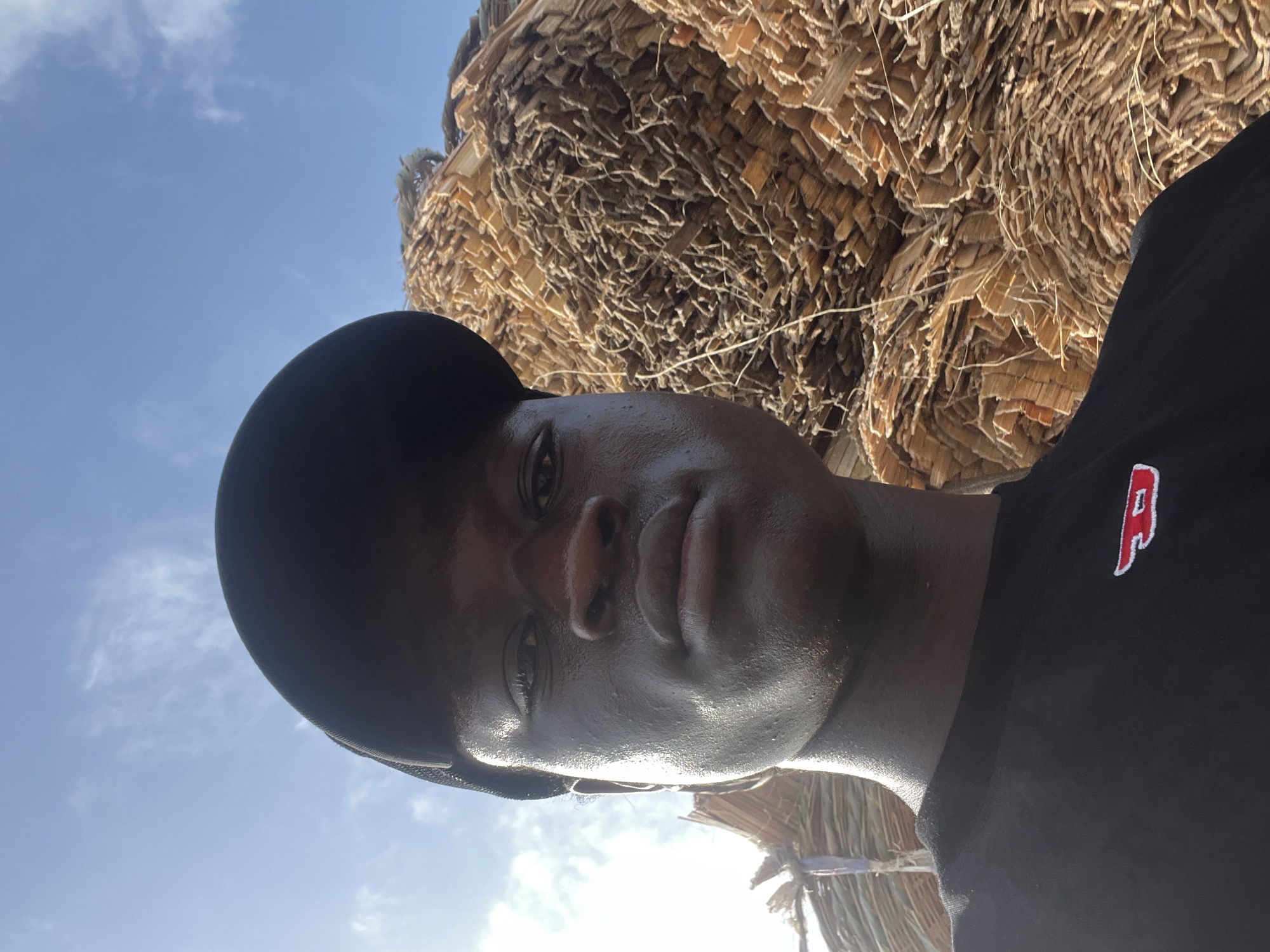 A man is seen in a close-up, low-angle shot. He has dark skin, a black baseball cap, and is wearing a black shirt with a red "P" logo. His face appears sweaty and reflective in the sunlight. Behind him is a backdrop of brown, thatched material, suggesting a roof or structure made from natural fibers. The sky is visible in the upper left of the frame, a pale blue with faint clouds. Given the prompt context, the location is presumed to be Badagry, Nigeria.