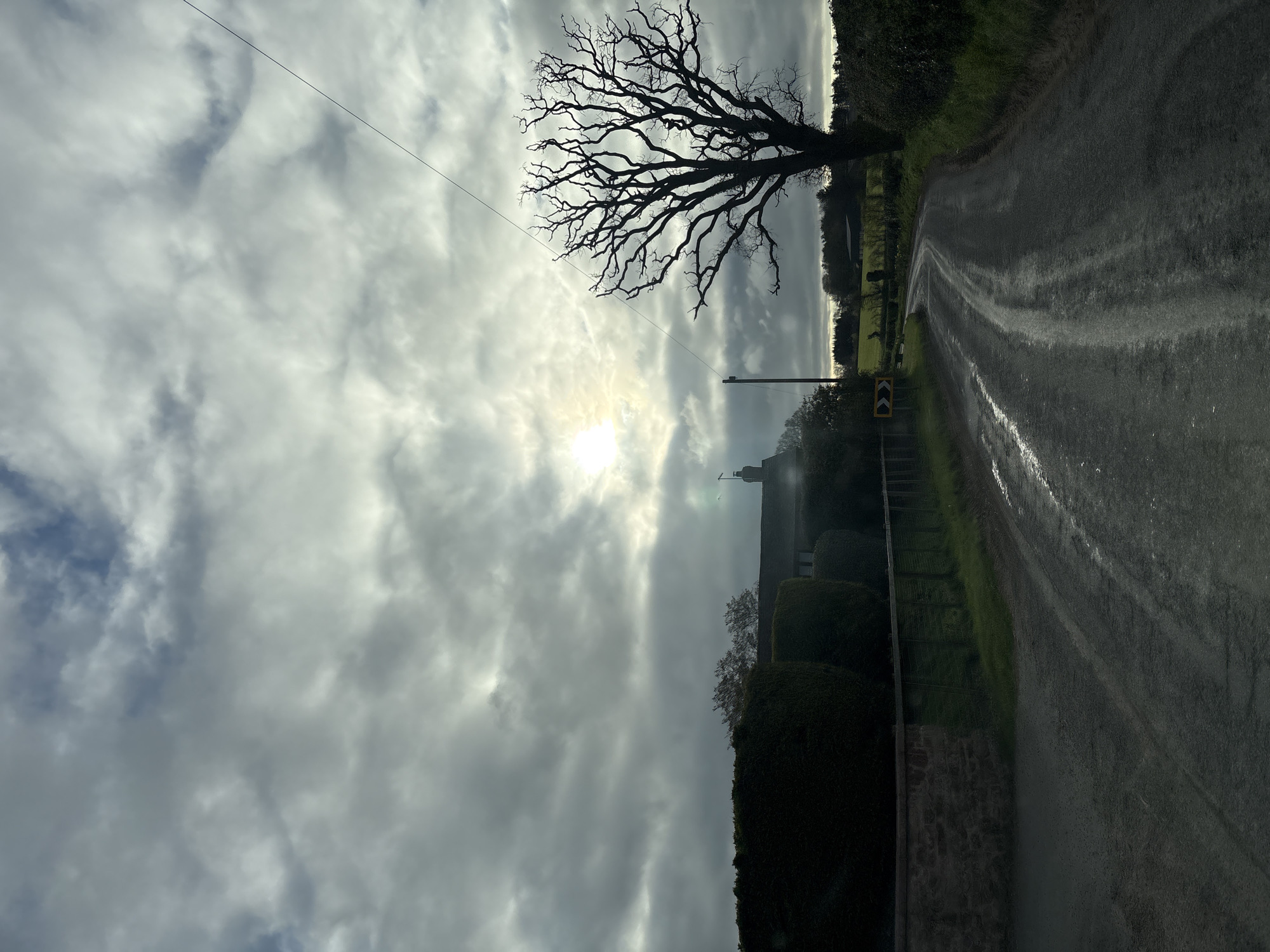 The image depicts a rural scene near Ruyton-XI-Towns, United Kingdom. A narrow, wet asphalt road curves to the right, marked by a yellow directional sign. A bare tree with intricate branches stands silhouetted against a cloudy sky, with the sun obscured by the clouds. A telephone pole is visible near the tree. A stone wall and hedges line the edge of the road. In the distance, a building, possibly a church, is partially obscured by trees. The sky is filled with gray and white clouds, creating a diffused light across the scene.