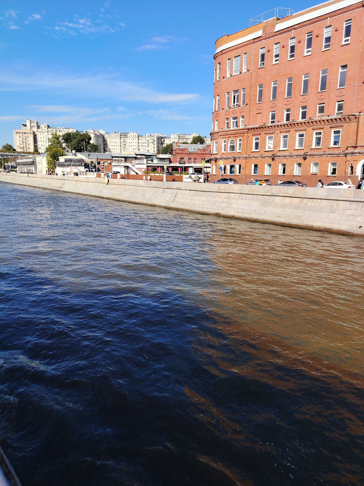 The image depicts a waterfront scene in Moscow, Russia, on a sunny day with a clear blue sky and scattered clouds. In the foreground, a wide river occupies the lower half of the frame, with a distinct color gradient from dark blue on the left to a murky brown on the right, suggesting different water conditions or reflections. The riverbank is lined with a concrete embankment.
Along the embankment, a row of buildings is visible. The most prominent building on the right is a large, multi-story structure made of red brick, with numerous white-framed windows. To its left, there are several other buildings, including some lighter-colored, multi-story apartment buildings in the background. Several cars are parked along the embankment, and a few individuals are present, walking or standing near the water's edge. Green trees are interspersed among the buildings. The overall atmosphere is urban and appears to be a developed area along the river.