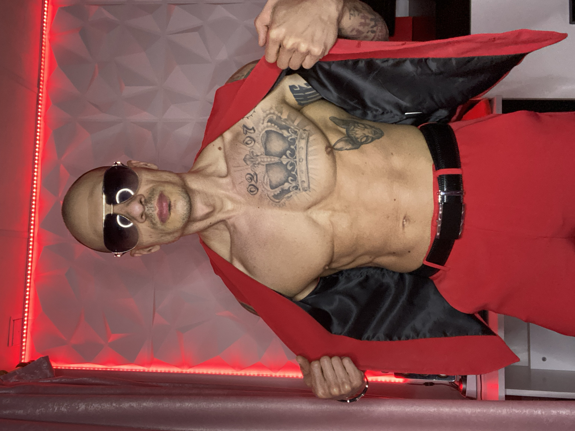 A bald, muscular man with sunglasses stands shirtless, wearing red pants and a red jacket with a black lining. He has multiple tattoos, including one of a crown with the number "2020" on his chest. The background consists of a white, geometric wall panel and red LED lighting. The location in the city of Pleven, Bulgaria, is not apparent in the image.