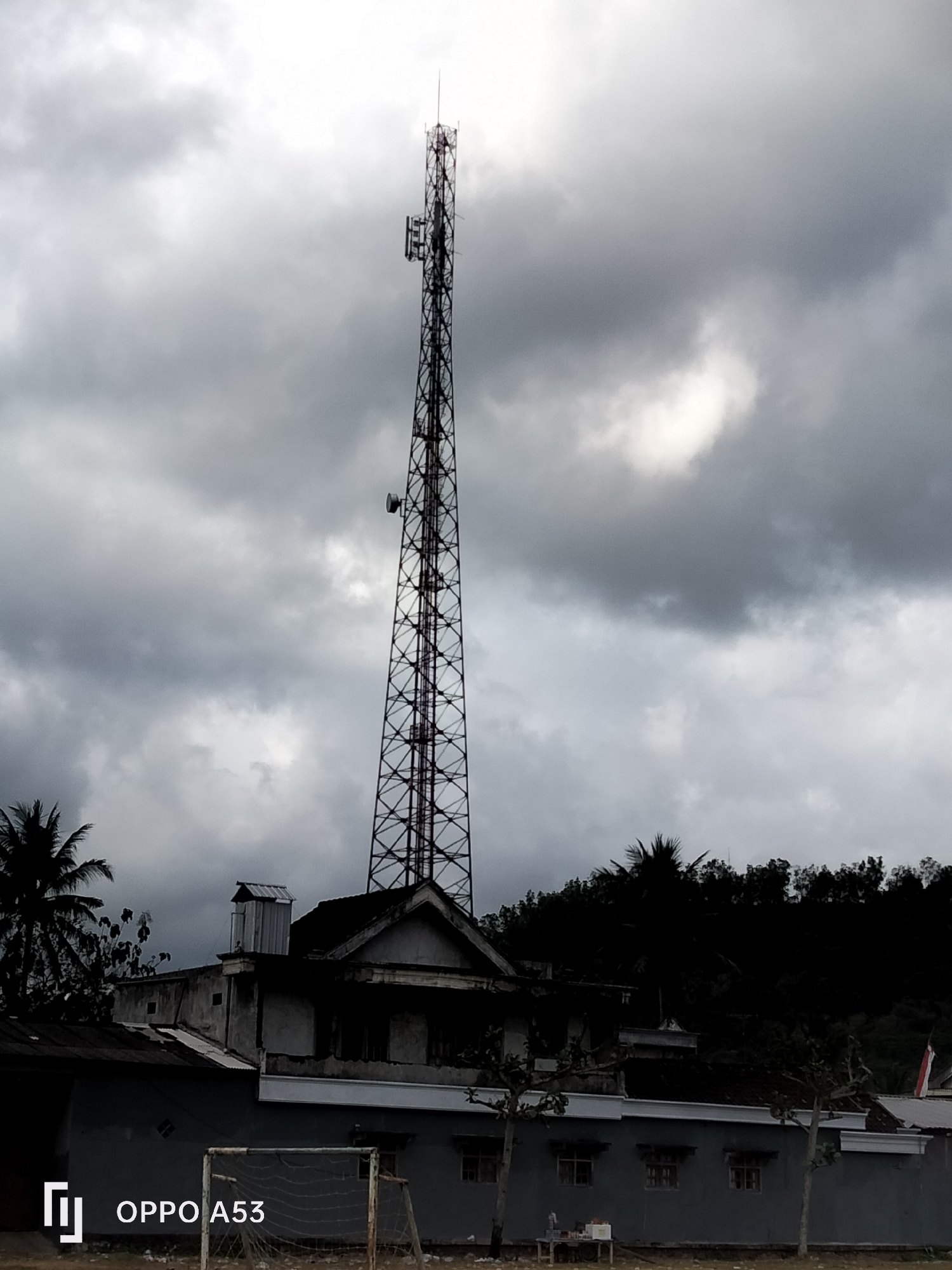 In Bendoagung, Indonesia, a tall, lattice-style telecommunications tower dominates the skyline under a dramatic, cloudy sky. In the foreground, a building with a dark, sloped roof and visible windows is situated below the tower. A sports net, possibly for soccer, is visible on the left side of the frame, partially obscured by the building. The overall scene suggests a somewhat overcast day in a semi-urban or rural setting.