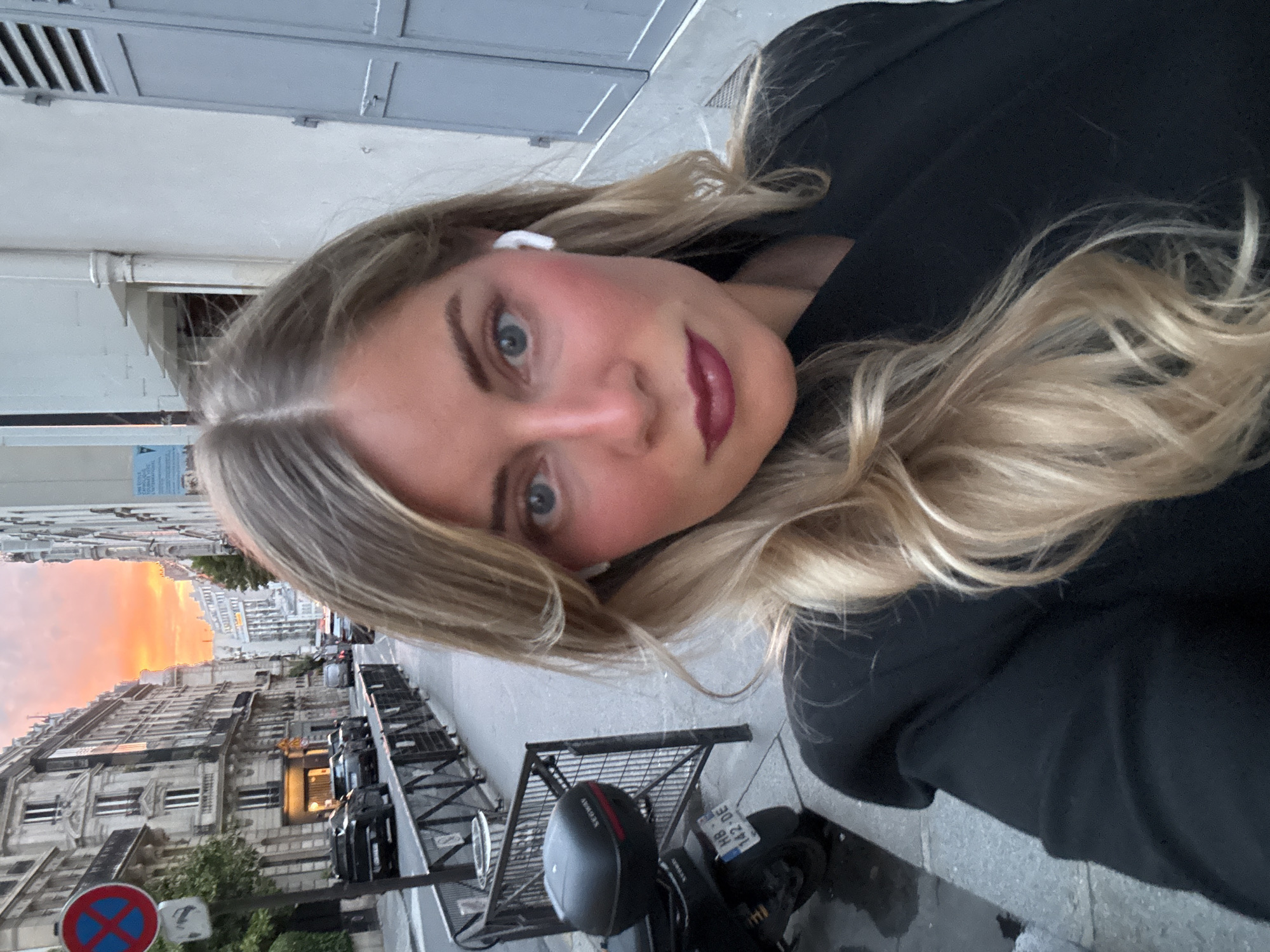 A woman with long, blonde hair and wearing wireless earbuds poses for a selfie. She is wearing a dark-colored jacket. The background shows a street in Paris, France, with traditional Parisian buildings visible along the street. A red and blue "no stopping" sign is on a pole, and parked cars line the street. A motorbike is parked nearby. The sky in the distance is a warm orange and red color, indicating either sunrise or sunset. The scene is urban and appears to be early morning or late evening.