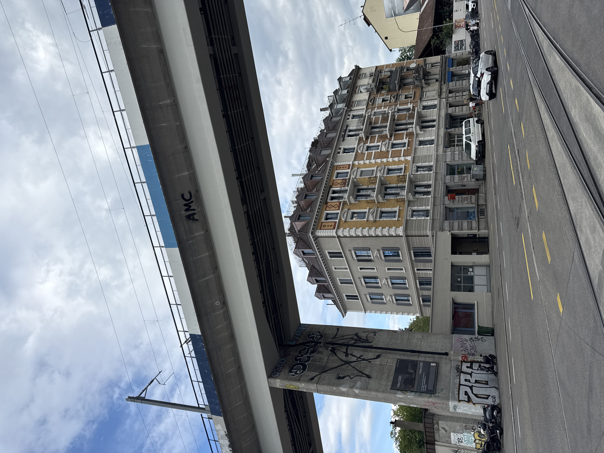 This is a vertical, low-angle shot of an urban scene in Zurich, Switzerland, under a cloudy sky. The foreground is dominated by the underside of a concrete overpass, featuring graffiti. One section of the overpass has "AMC" spray-painted in black lettering, while another pillar below shows larger, more complex graffiti tags, including "OSLO AL LUMPE", "ACAB", and "SM Avaris", along with a poster for "Volvo ES90" and "Volvo EX90".
Beyond the overpass, a multi-story building with a yellow facade and numerous windows stands tall. The building's architecture suggests an older European style, with decorative elements and balconies. The street level shows tram tracks running parallel to the sidewalk, where parked motorcycles and cars are visible. A white van and a white car are parked in front of the building. The scene appears to be a typical street in a city, likely during daylight hours, given the ambient light. The presence of tram tracks, the architecture, and the graffiti point to a bustling urban environment.