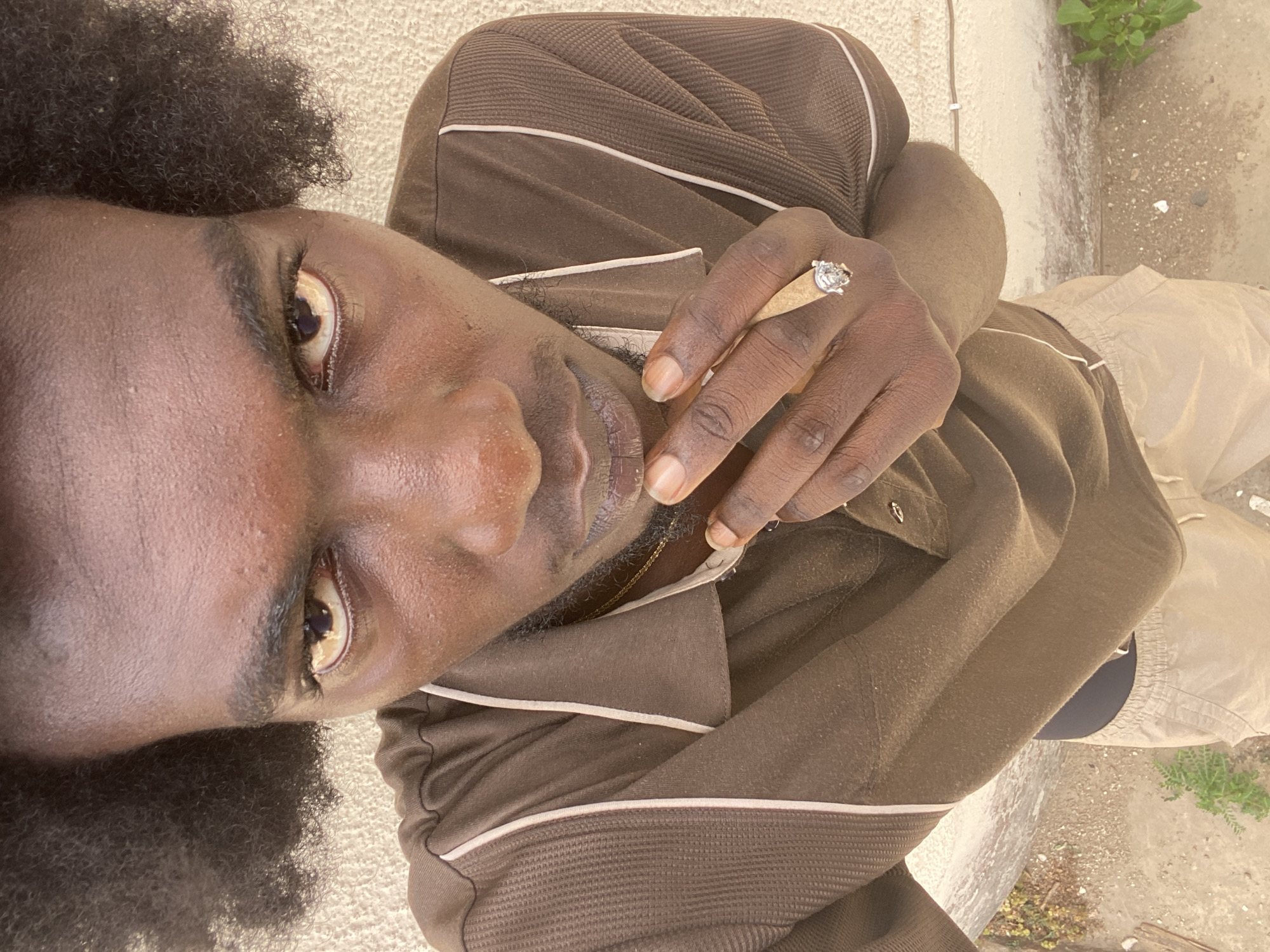 A dark-skinned man with tightly curled black hair is holding a lit marijuana cigarette between his fingers. He is wearing a brown long-sleeved shirt with white stripes and a ring on his left hand. He appears to be standing outside on a concrete surface. The location cannot be determined to be Lagos, Nigeria from visual information.