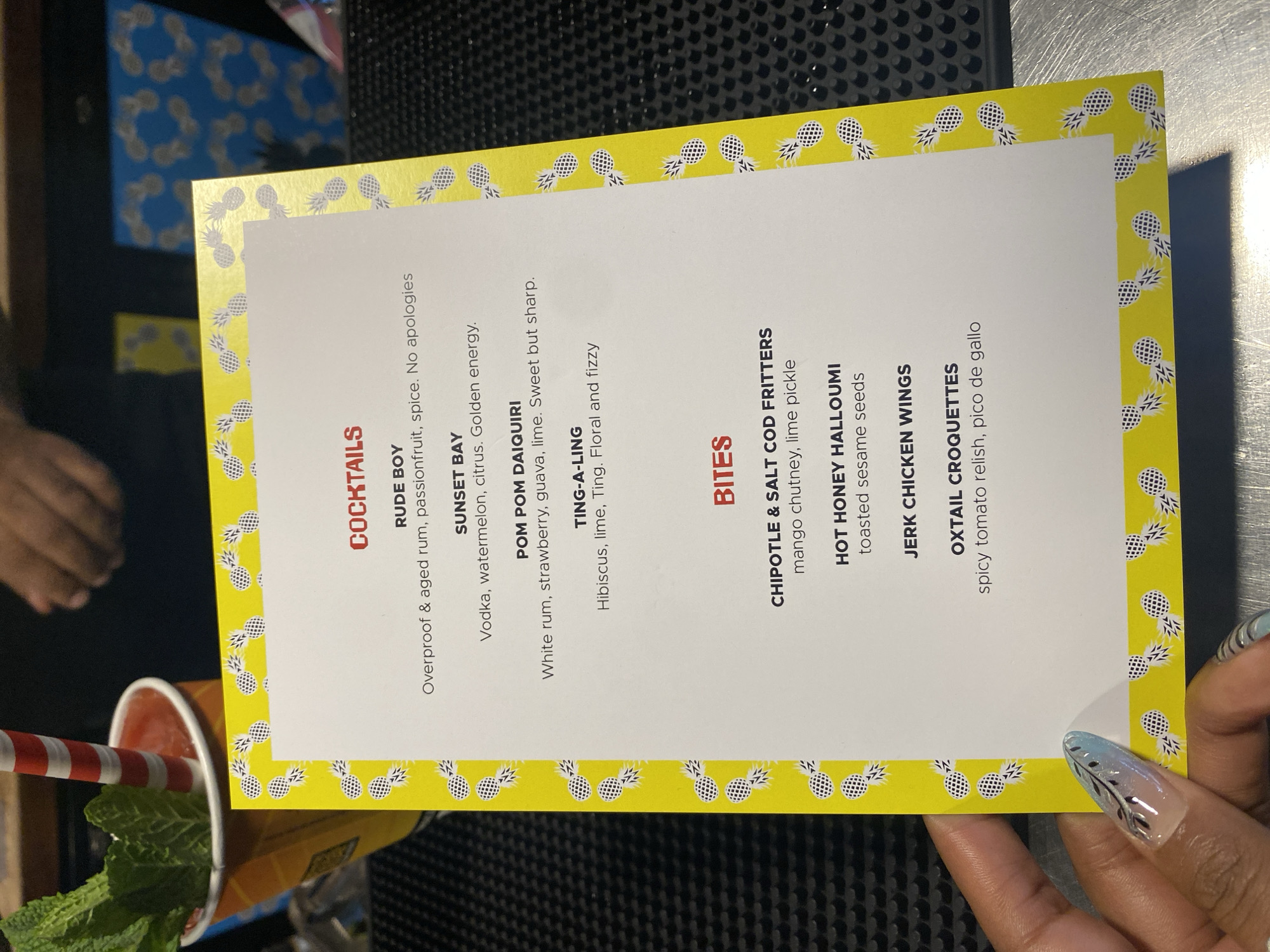 Here is a description of the image:
The image shows a menu card with a pineapple border, listing cocktails and bites. The cocktail options are "RUDE BOY" (overproof & aged rum, passionfruit, spice), "SUNSET BAY" (vodka, watermelon, citrus), "POM POM DAIQUIRI" (white rum, strawberry, guava, lime), and "TING-A-LING" (hibiscus, lime, Ting). The bites include "CHIPOTLE & SALT COD FRITTERS" (mango chutney, lime pickle), "HOT HONEY HALLOUMI" (toasted sesame seeds), "JERK CHICKEN WINGS", and "OXTAIL CROQUETTES" (spicy tomato relish, pico de gallo).
Beside the menu is a drink with a striped straw and mint garnish. A hand with decorated nails is holding the menu.
It is not possible to determine the exact location in London from this image alone.