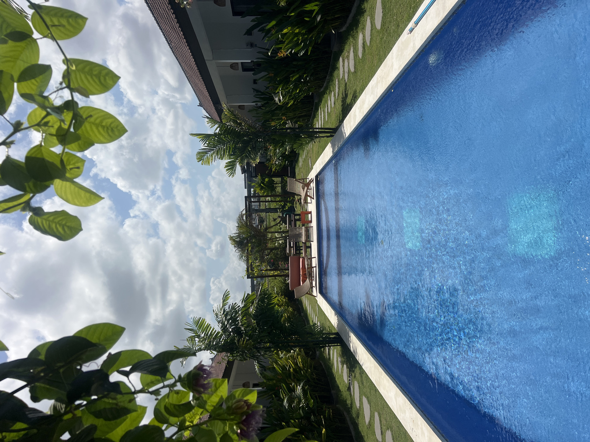 This is a vertical, eye-level, outdoor shot of a resort swimming pool area in North Kuta, Indonesia. The pool, filled with vibrant blue water, dominates the foreground and extends into the midground. The pool is framed by a light-colored coping and bordered by a grassy lawn. Stepping stones are laid out on the grass along the edge of the pool. To the left and right of the pool are two single-story buildings with dark tiled roofs, typical of Balinese architecture. Lush green foliage, including palm trees and other tropical plants, flanks the buildings and the pool area. In the midground, on the right side of the pool, are several lounge chairs and a small table, suggesting a place for relaxation. Further in the background, a wooden pergola with some climbing plants is visible. The sky above is a bright blue with scattered, fluffy white clouds. The overall impression is one of a tranquil resort environment.