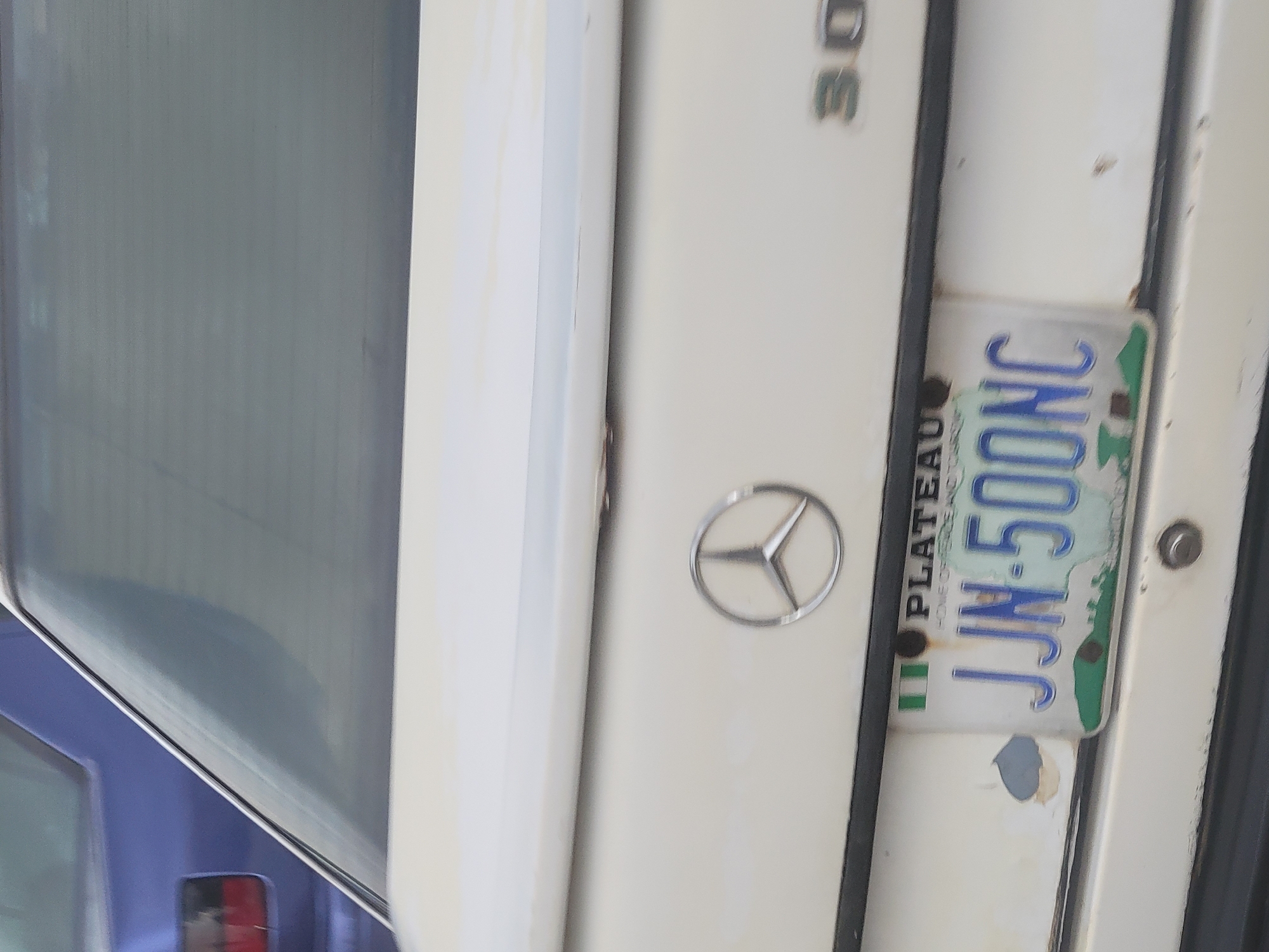 A partially visible white Mercedes-Benz vehicle, with its rear-end emblem and “30” visible on the trunk, features a Nigeria Plateau state license plate with the registration number JJN-500NC. The car's paint shows signs of wear. The vehicle is photographed in an unspecified location in the city of Gura Top, Nigeria.