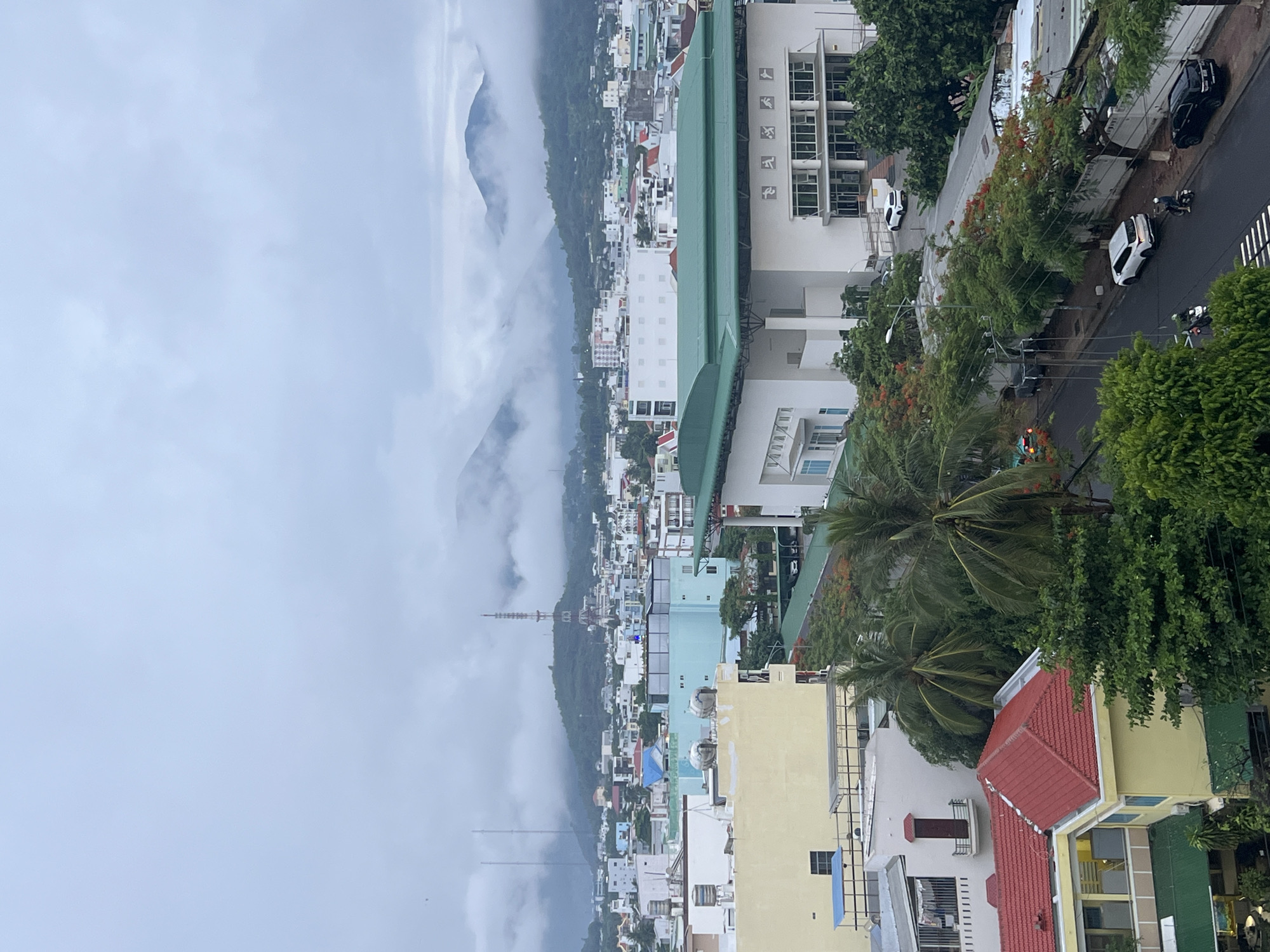 This is an outdoor, elevated, partially obscured view of Nha Trang city in Vietnam. Dense fog and clouds conceal the left half of the image, partially obscuring a mountain range. Below the mountain, a dense urban landscape stretches towards the right side of the frame. The right half of the image is dominated by buildings, streets, and vegetation. Buildings vary in height and color. The building closest to the view has a red roof. Streets are lined with trees and parked vehicles, including cars and scooters. Power lines are visible. It appears to be daytime.