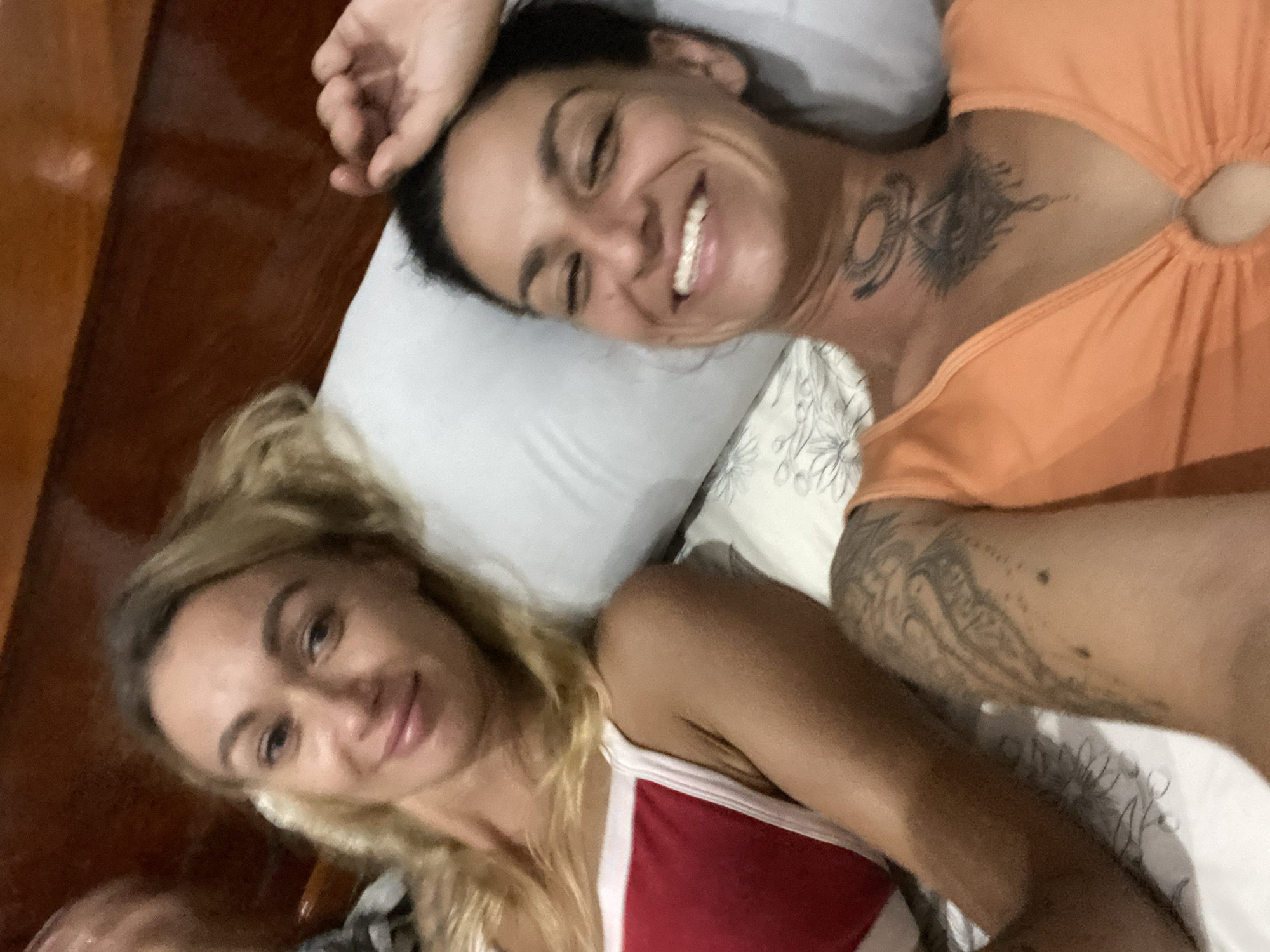 Two women are lying in bed. The woman on the left has blonde hair and is wearing a red and white top. The woman on the right has dark hair and is wearing an orange top. She has tattoos on her neck and arm. The image appears to have been taken indoors in a bedroom, possibly in a hotel. The location of Mũi Né, Vietnam is not determinable from the image.