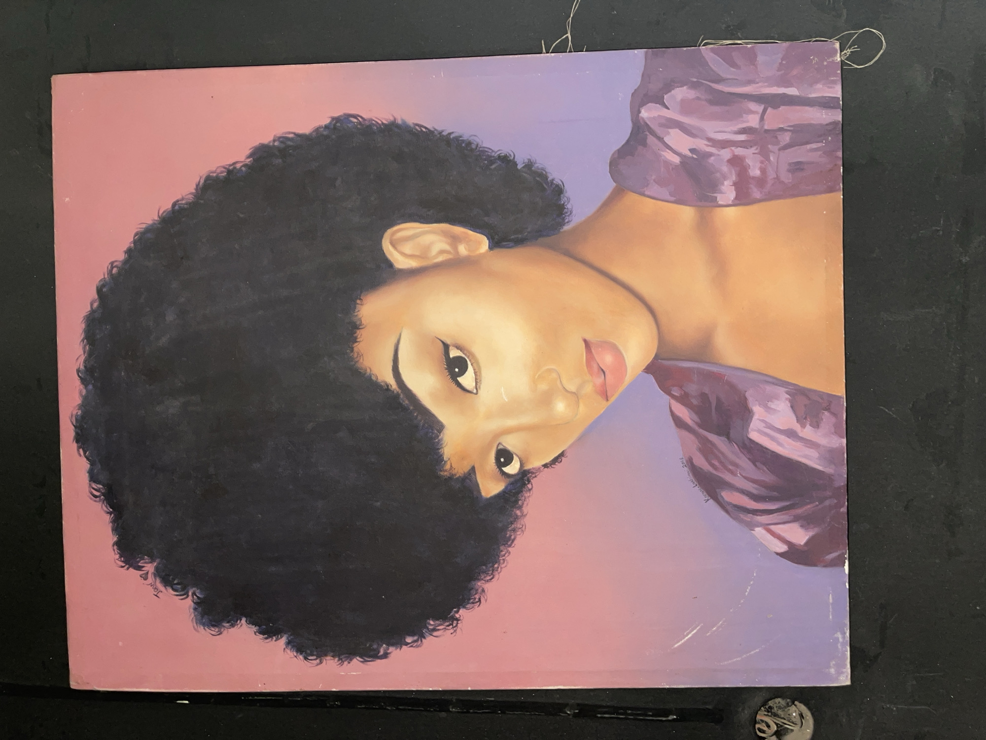 An oil painting depicting a dark-skinned woman with an afro, wearing a purple garment. The background is a gradient of pink and purple. The painting is signed "Kwarshak Jokthan 2018." There is no indication of the location in the city of Jos, Nigeria. The image appears to be a photograph of the painting.