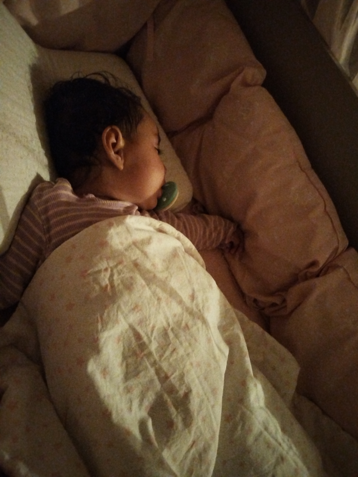 An overhead shot shows a baby sleeping on its side, swaddled in a patterned blanket. The baby is wearing a striped shirt and has a pacifier in its mouth. The baby is lying on a bed or couch with pillows. The scene appears to be indoors, in a dimly lit room.