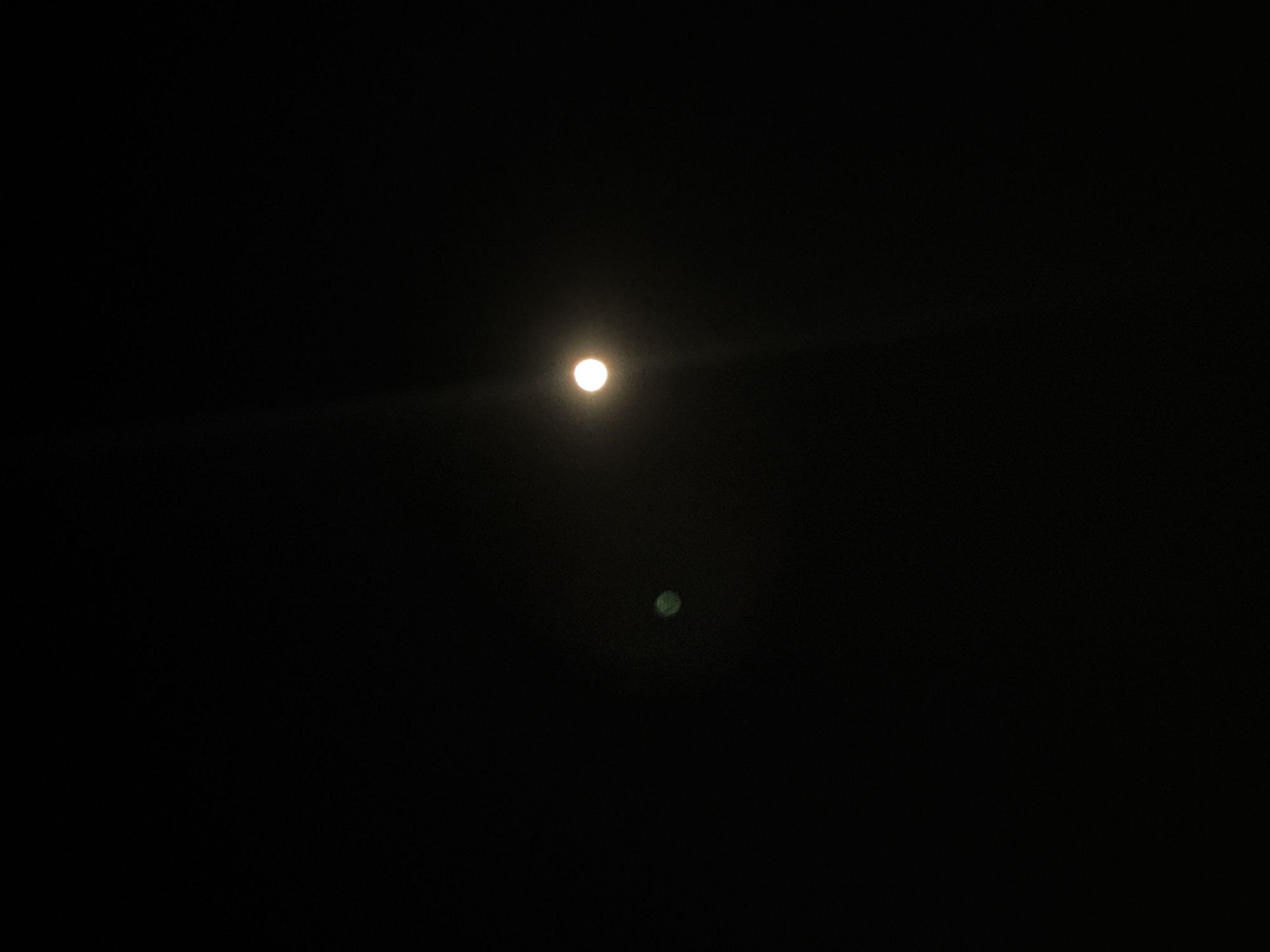 The image is a vertical, medium shot of a dark night sky with two celestial bodies visible. The primary object is a bright, white, circular disc emitting a soft glow and a faint light flare extending upwards. To the left and slightly lower than the main object, there is a smaller, blurry, green circular object. The background is entirely black, indicating a lack of other visual elements or ambient light. The image's composition suggests it was taken from a ground-level perspective looking upwards. Without additional information or context, it is impossible to determine the specific location within the city of Opoczno, Poland, or any specific details about people, actions, or interactions. The scene is purely astronomical in nature.