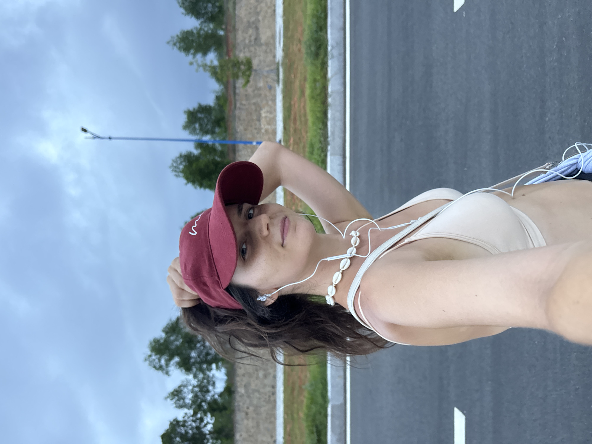 A woman is captured in a selfie outdoors, holding her ponytail and wearing a red baseball cap, a light-colored sports bra, and a necklace of shells. She has white earbuds in her ears. The background consists of a road with white lines, a grassy embankment, a stone wall, trees, and a cloudy sky. A blue lamppost is visible in the distance. The scene appears to be in Mui Ne, Vietnam, a coastal city known for its beaches and sand dunes.