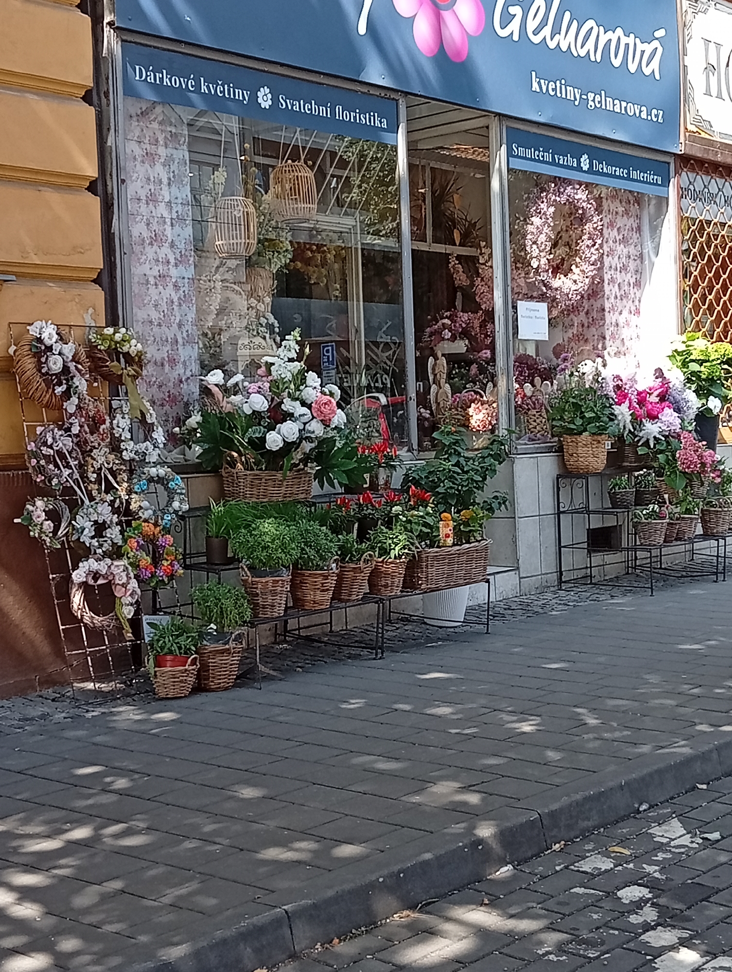 The image shows a flower shop with the name "Gelvarova" above the entrance. The shop displays a variety of floral arrangements, wreaths, and potted plants on the sidewalk. The building appears to be located in a city with a cobblestone sidewalk and a yellow-toned building facade. The shop's exterior features large glass windows showcasing the interior, which is filled with flowers and decorations. The exterior display includes a metal rack holding numerous floral wreaths and a selection of potted plants arranged on metal shelves. The scene is lit by natural daylight, casting shadows from nearby trees or structures onto the pavement. The overall impression is that of a well-stocked, inviting flower shop in a European city. The specific location in Brno, Czechia, is confirmed by the signage on the shop, which features Czech text.