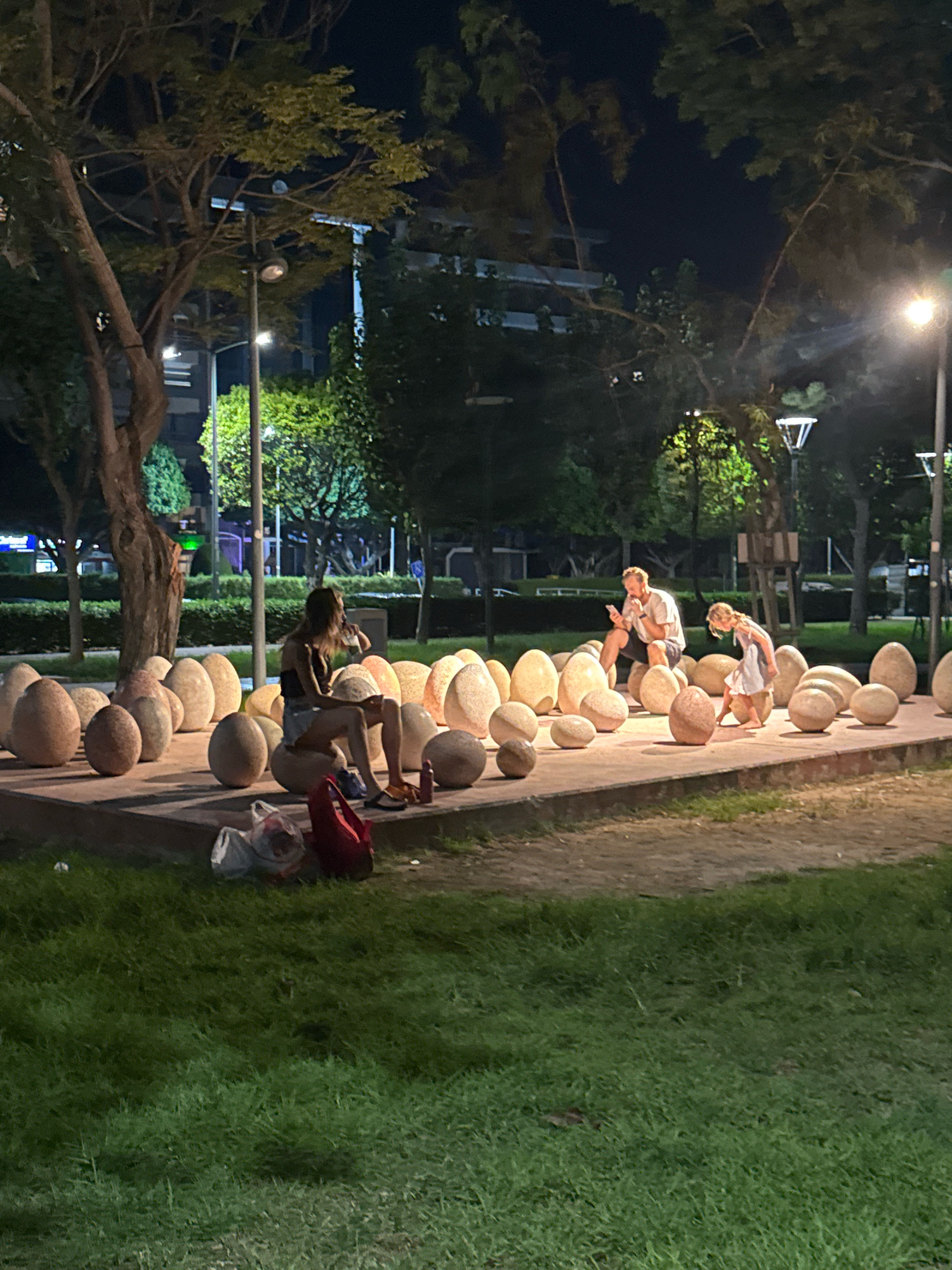 In the city of Limassol, Cyprus, at night, an outdoor seating area is illuminated by streetlights. The area is adorned with numerous large, smooth, egg-shaped stones arranged in a display. Three people are present. A woman sits on one of the stones, holding a drink. A man sits on another stone nearby, looking at his phone. A child stands between them, interacting with the stones. In the foreground, a patch of green grass is visible. Bags and a red tote bag are placed on the ground in front of the seating area. The background shows trees, buildings, and more streetlights.