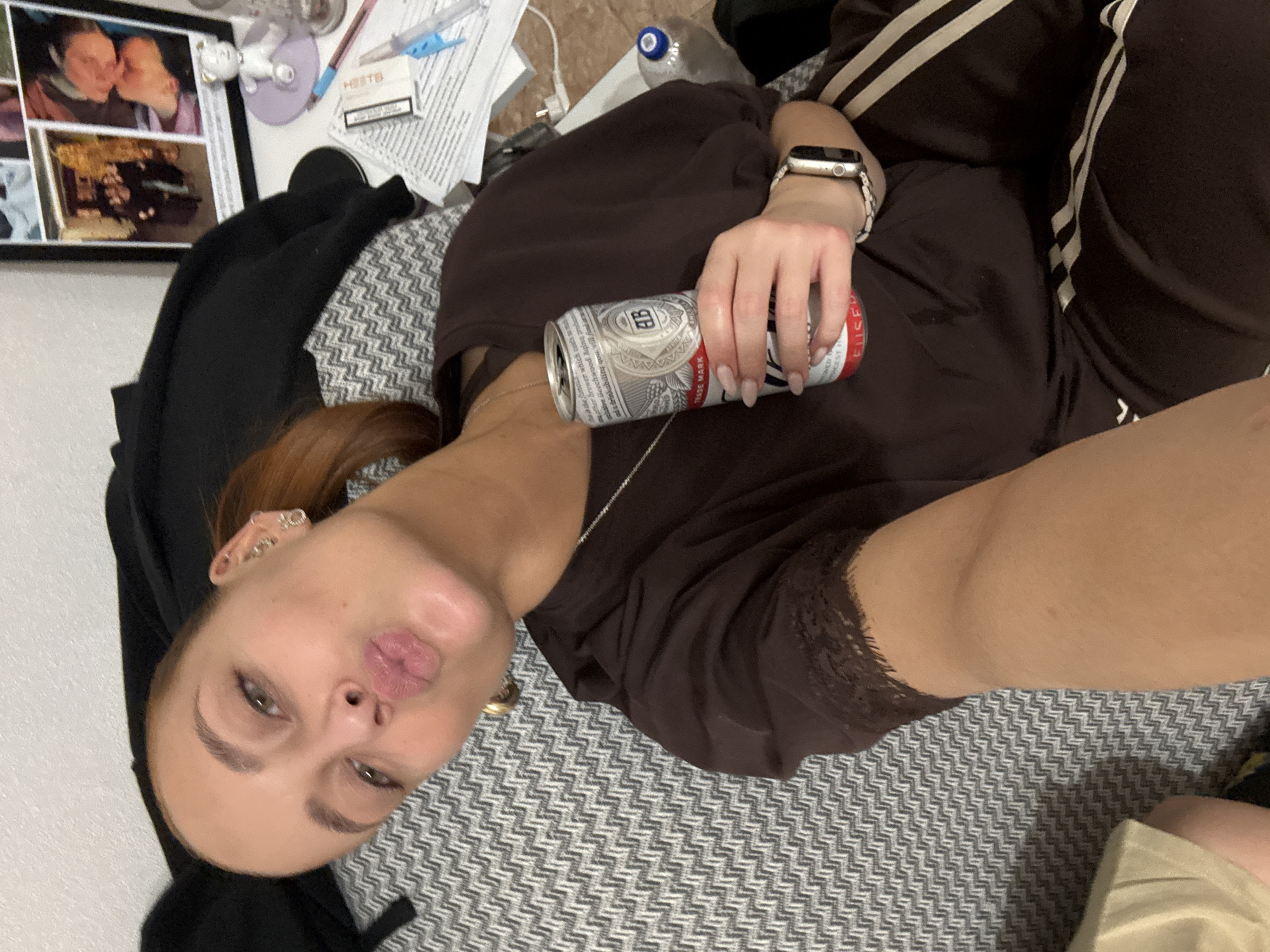 A woman lies on a grey and white patterned surface with her head toward the bottom left of the frame. She has fair skin, brown hair pulled back in a ponytail, and wears multiple earrings in her left ear. Her lips are puckered. She is dressed in a brown top with lace detailing around the neckline and brown pants with cream stripes down the sides. She holds a can of Budweiser beer in her right hand, on which she wears a smart watch.
To the left of her head are a framed photo collage, papers, cigarettes, and other items. A black garment is draped nearby. A water bottle and other objects are also visible. The location is likely indoors. There is no indication the image was taken in Chaikovsky, Russia.