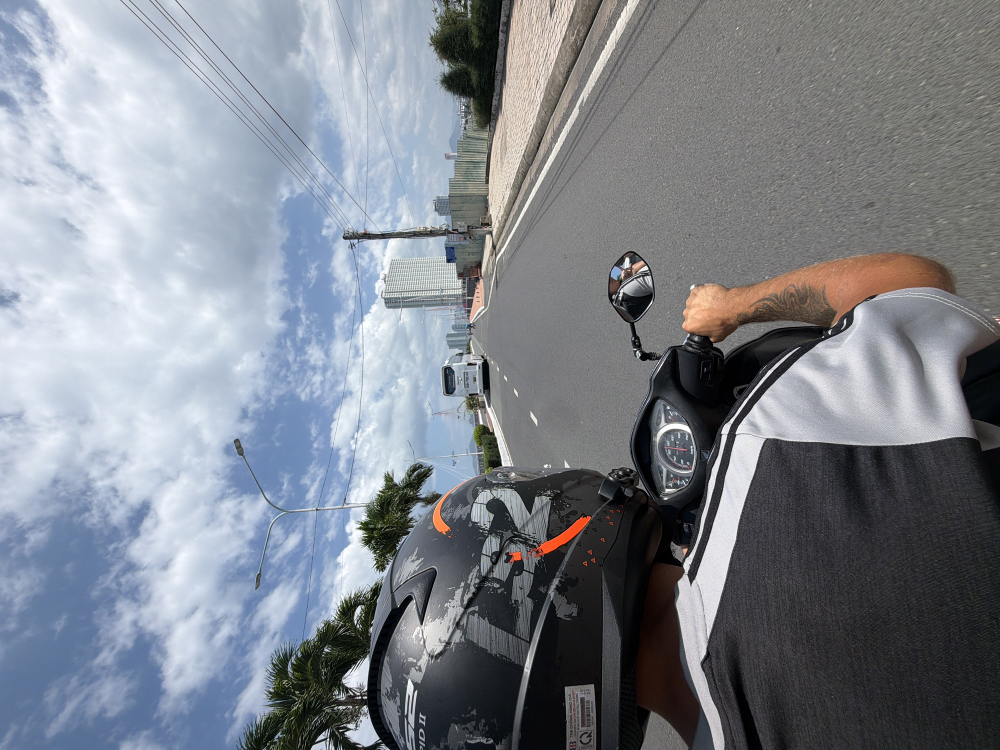 The image presents a first-person perspective from an individual riding a motorcycle or scooter along an urban road in Nha Trang Ward, Vietnam. The view is directed forward, slightly angled towards the left. In the foreground, the back of a black LS2 RAPID II helmet with striking grey and orange graphic designs is prominent. A label on the helmet includes the text "TEM CHONG HANG GIA" (anti-counterfeit stamp) and a QR code. The rider's right arm, featuring a black tattoo, is visible gripping the scooter's handlebar, while a portion of their torso, wearing a black shirt with white side stripes, is also seen.
The scooter's dashboard, showing a speedometer and other gauges, is partially visible, and the right rearview mirror reflects part of the rider's arm and shirt. The asphalt road ahead has clear white lane markings. Along the right side of the road, there's a brick-paved sidewalk lined with green trees and bushes, leading towards a cityscape with tall, modern buildings in the distance, some under construction with visible scaffolding. A white bus and another vehicle are observed further down the road.
To the left, palm trees and utility poles with power lines are silhouetted against a bright, cloudy sky. Modern streetlights with two arms extend overhead. The scene depicts a clear, sunny day with scattered clouds, showcasing a dynamic urban environment with ongoing development in Nha Trang, Vietnam.