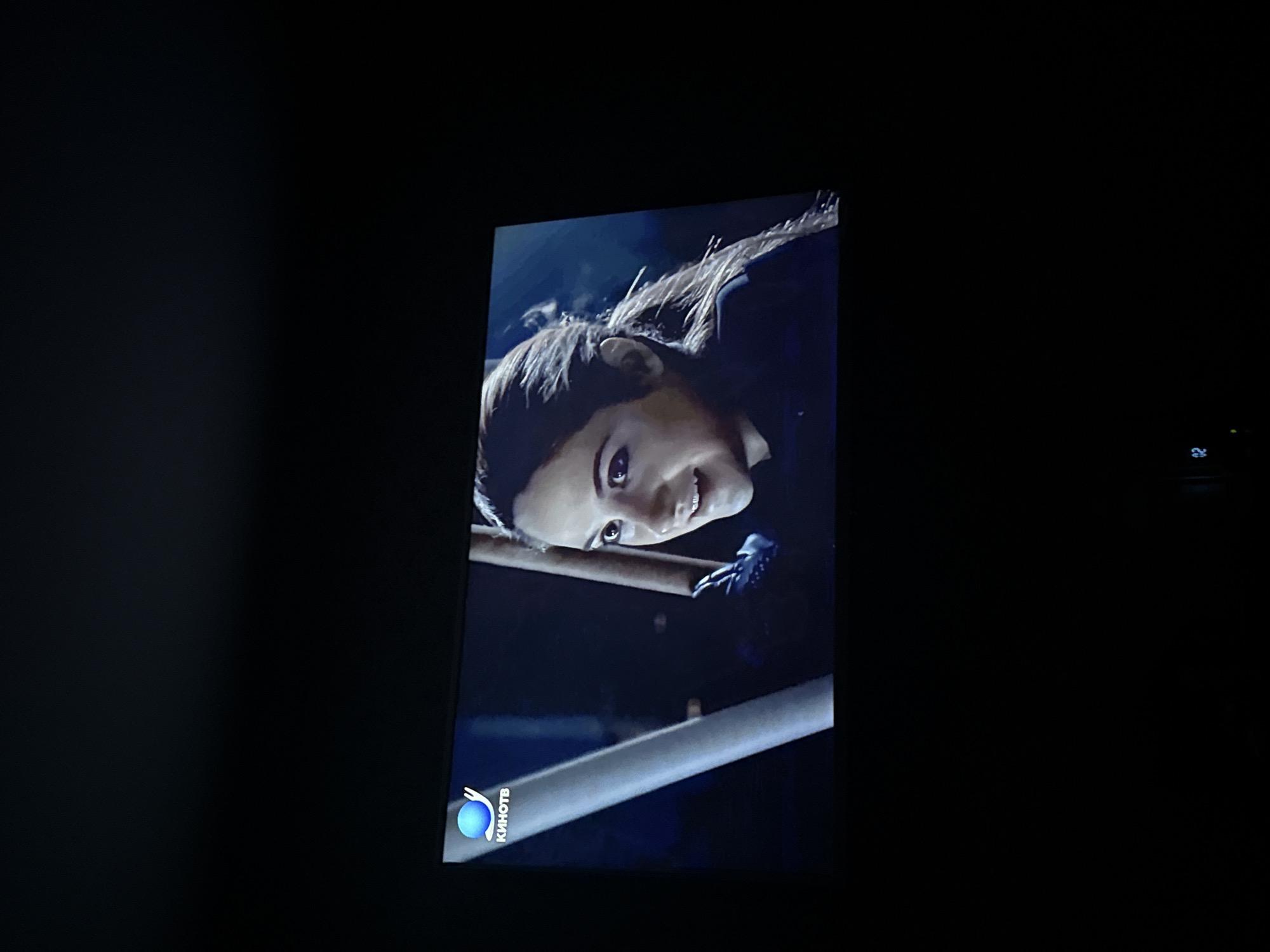 The image shows a close-up of a woman's face on a TV screen. She appears to be holding onto a metal bar or railing. The image is dark, with the woman's face and the railing the brightest parts of the scene. The television screen also displays a "Kinotv" logo in the lower left corner. No information about Mytishchi, Russia can be inferred from the image.