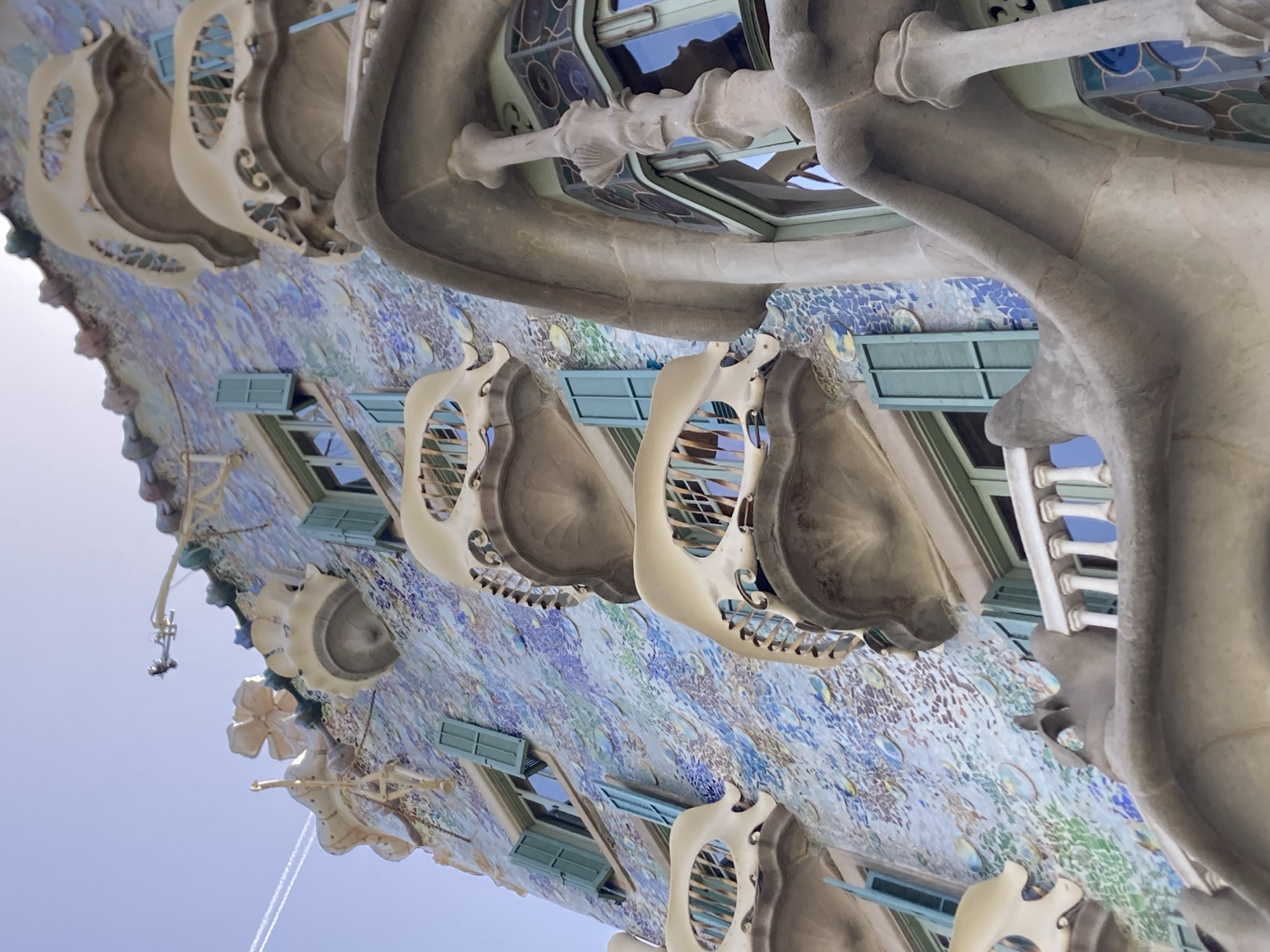This is a close-up, low-angle shot of the exterior of a building with a distinctive, organic architectural style. The building's facade is adorned with a vibrant mosaic of blue, green, and white tiles, creating a shimmering, textured surface. Several windows with light blue shutters are visible.
Emerging from the facade are large, bone-like, cream-colored structures that appear to be balconies or decorative elements. These elements have an intricate, skeletal design with openwork sections. Above these, more of the building's unique architecture is visible, featuring curved, stone-like formations and stained glass windows with circular patterns. The overall impression is of an unusual, almost fantastical structure.
The image is taken from below, looking upwards towards the sky, which is a pale, hazy blue, suggesting it might be a bright but overcast or hazy day, or perhaps early morning or late afternoon light. There is no visible text in the image. The focus is entirely on the unique architectural details and the interplay of light and shadow on the building's surface. The context provided places the location as Barcelona, Spain, which is consistent with the architectural style that evokes Gaudí's work.