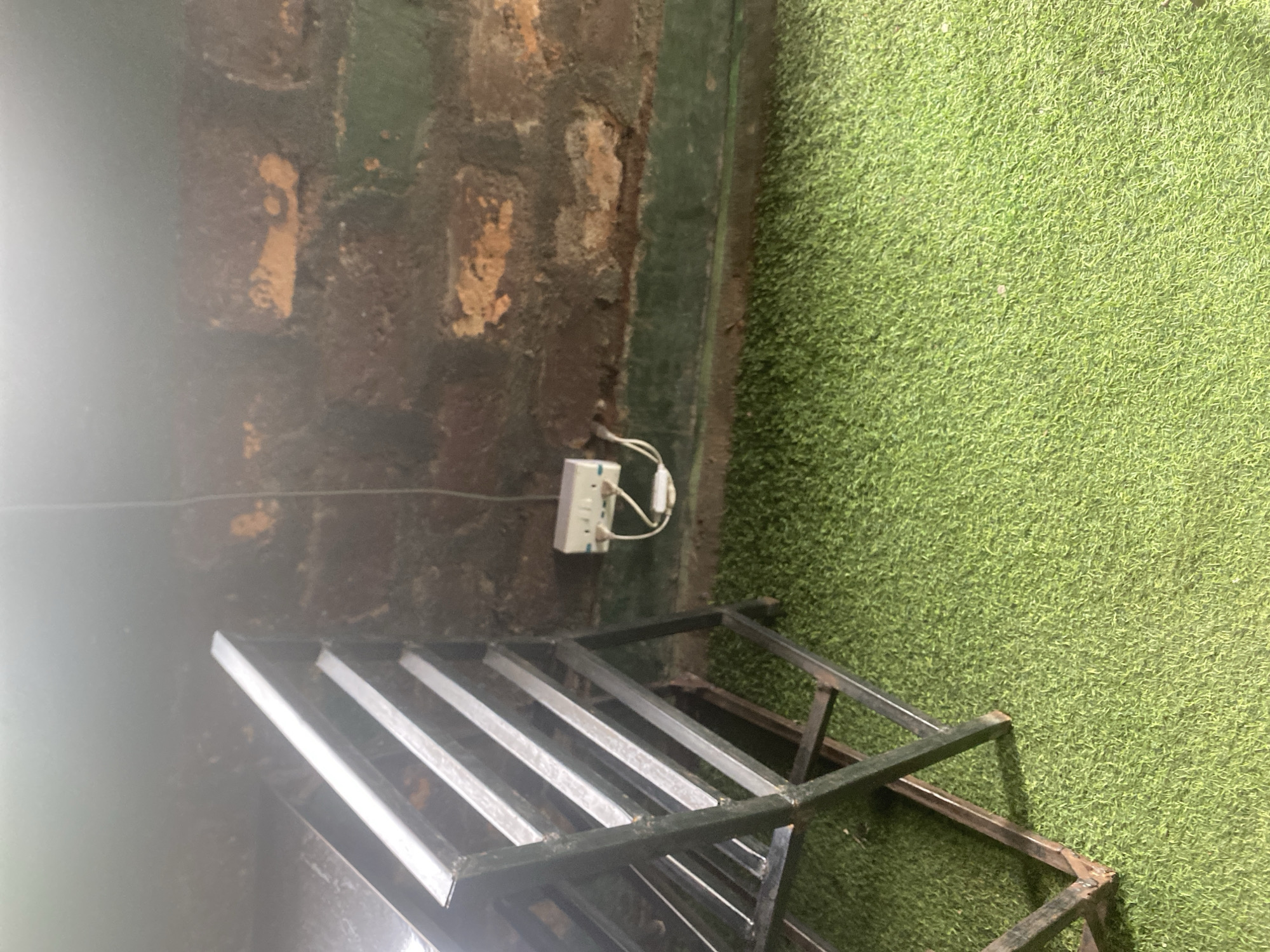 The image depicts an indoor scene with a brick wall on the left and artificial turf on the right. A white electrical power strip with two plugs connected to it is mounted on the brick wall. In the foreground, a metal shelf unit with several horizontal bars is visible. The lighting in the image is dim, with a bright light source or glare on the left side, obscuring some details. The overall impression is that of an utilitarian space, possibly a storage area or a workshop, given the presence of the shelving unit and electrical outlet. There is no indication of time of day, weather, or specific emotions. No text is visible in the image.