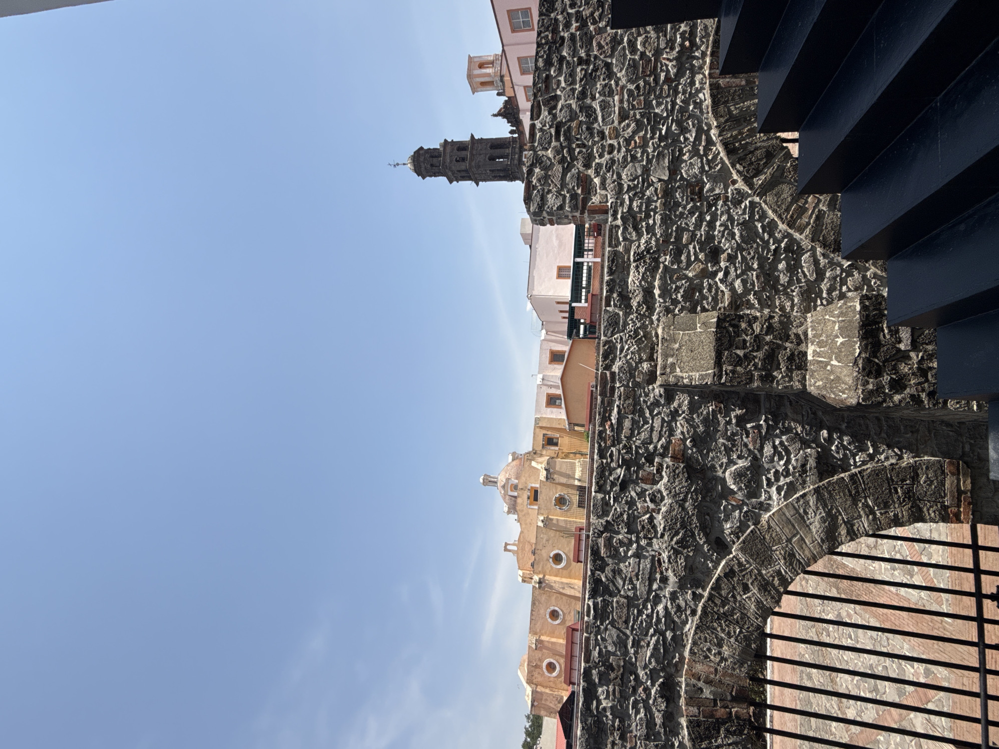 The image, oriented on its side, captures a historical architectural scene under a clear blue sky.
In the foreground and running along the right side is a prominent, weathered stone wall, constructed from large, irregularly shaped dark grey and brown rocks. This rough texture suggests an ancient or colonial structure. Partially obscuring the upper right portion of the wall are several dark, angular, horizontal beams or steps, which appear to be part of a more modern architectural element, casting sharp shadows. At the bottom right, an arched opening within the stone wall is visible, fronted by dark, vertical iron bars or a gate.
Beyond the stone wall, against a bright, cloudless sky, a cluster of old buildings forms the background. The most distinct is a tall, ornate church tower, dark in color, featuring multiple tiered sections and topped with a cross. To its right, another historic building with a yellowish-ochre facade, a domed roof, and several circular windows can be seen. Various other light-colored structures, including pink, white, and orange buildings, are scattered throughout the mid-ground.
The time of day is clearly bright daylight, evidenced by the brilliant blue sky and the crisp shadows. The weather is sunny and clear. No people or specific events are visible; the scene is static, highlighting the historical architecture. The architectural style, particularly the church and colonial buildings, strongly aligns with the specified location of Puebla, Mexico. There is no visible text in the image.