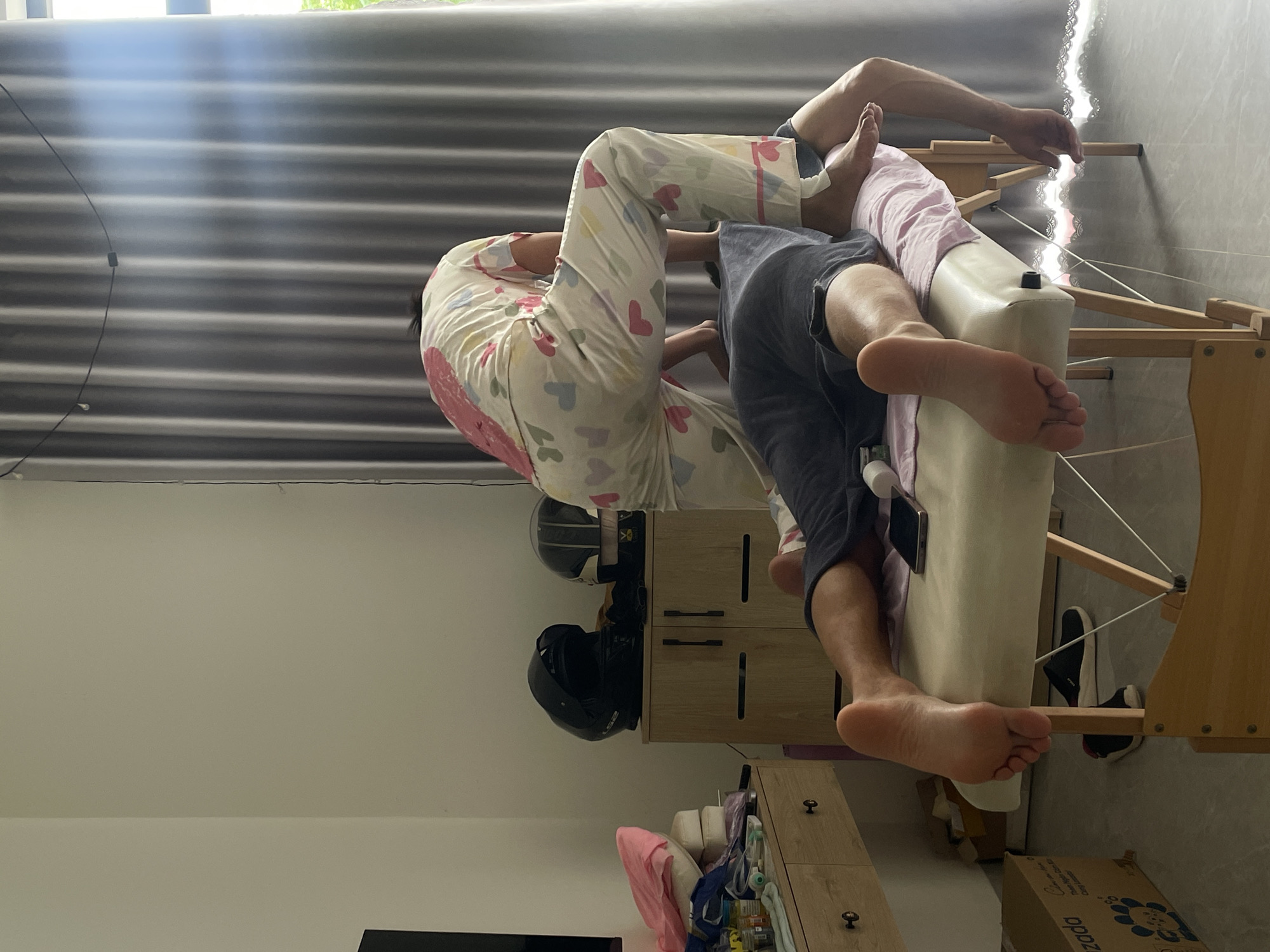 This image captures an indoor scene, likely a room within a home or a private spa, illuminated by bright natural daylight streaming through a large window. The central focus is a beige massage table with a wooden frame, where two individuals are engaged in what appears to be a massage or therapeutic session.
One person is lying prone on the massage table, positioned with their head towards the upper left and bare feet extended towards the lower right. They are wearing dark shorts, and a light pink garment covers part of their back and legs. A smartphone rests on the table near their left shoulder. Seated or kneeling at the head of the table, behind the first person, is the second individual, presumably the practitioner. This person is dressed in light-colored pajamas adorned with pink, blue, and yellow heart patterns.
In the background, on the right side of the room, there are two light wooden cabinets. The upper cabinet has two black motorcycle helmets placed on top, one of which faintly displays "LS2." The lower cabinet holds various items on its surface. A pair of dark sneakers with white soles are visible on the floor beneath the massage table. To the right of the sneakers, a partially visible cardboard box is present, bearing the text "Muzado" and a circular design. The window, located at the top of the frame, is covered by grey horizontal blinds. A thin black cable runs vertically along the wall near the far left edge of the window. The walls and ceiling are plain and light-colored. The setting, including the helmets, is consistent with the provided location context of Mũi Né, Vietnam.