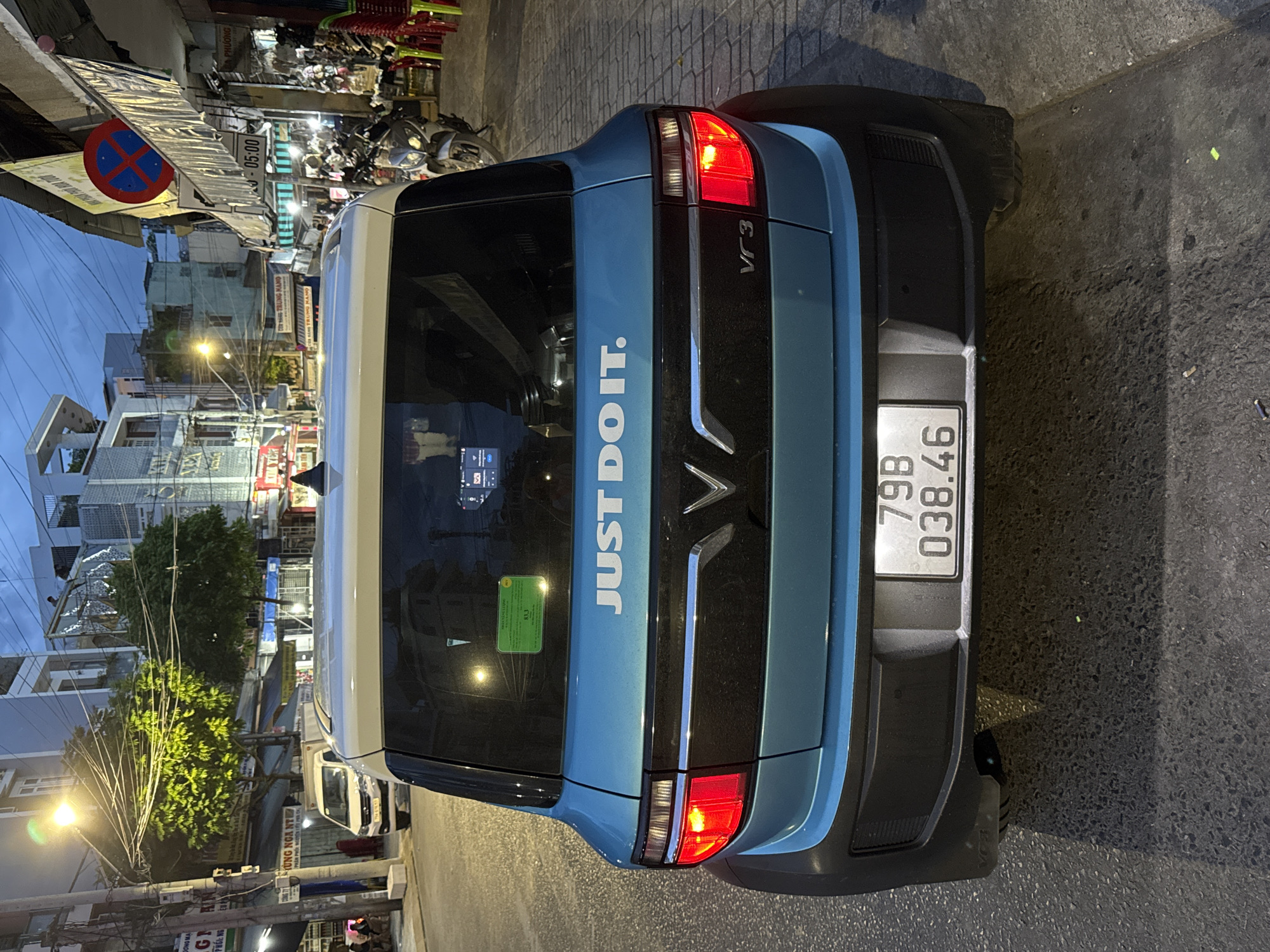 A rear-view, medium shot captures a blue and white VinFast VF 3 electric microcar parked on a street at dusk in Nha Trang, Vietnam. The car, emblazoned with "JUST DO IT." in white lettering on its rear, is the central focus. The license plate reads "79B 038.46". Streetlights cast a warm glow on the wet asphalt and the surrounding buildings, which appear to be shops and residences. A no-parking sign is visible in the background, suggesting a commercial or residential street. A few motorbikes are parked on the sidewalk, and red plastic chairs are stacked outside a storefront, indicating a commercial area. The sky is a dusky blue, with a few clouds visible. The overall scene depicts a typical urban streetscape in Vietnam during the evening.