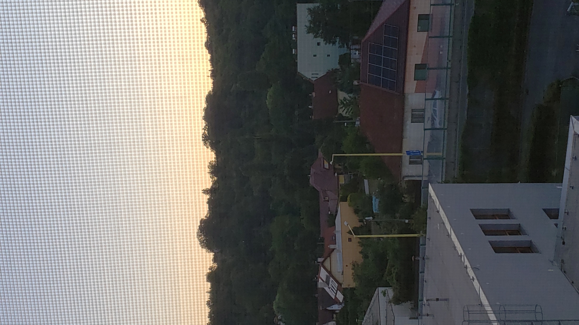 The image captures a view through a window screen, showing a residential area in Prešov, Slovakia, likely during the late afternoon or early evening. In the foreground, the flat roof of a light-colored building is visible, with a metal ladder structure attached to its side. Further back, a cluster of houses with red-tiled roofs is nestled among lush green trees. One house prominently features solar panels on its roof. A light blue, multi-story building stands to the right, with several windows visible. A yellow lamppost is positioned in front of some of the houses. The background is dominated by a densely wooded hill, silhouetted against a sky transitioning from pale blue to a warm, soft orange, suggesting sunset. The overall scene depicts a tranquil, suburban environment with a blend of residential buildings and natural landscape.