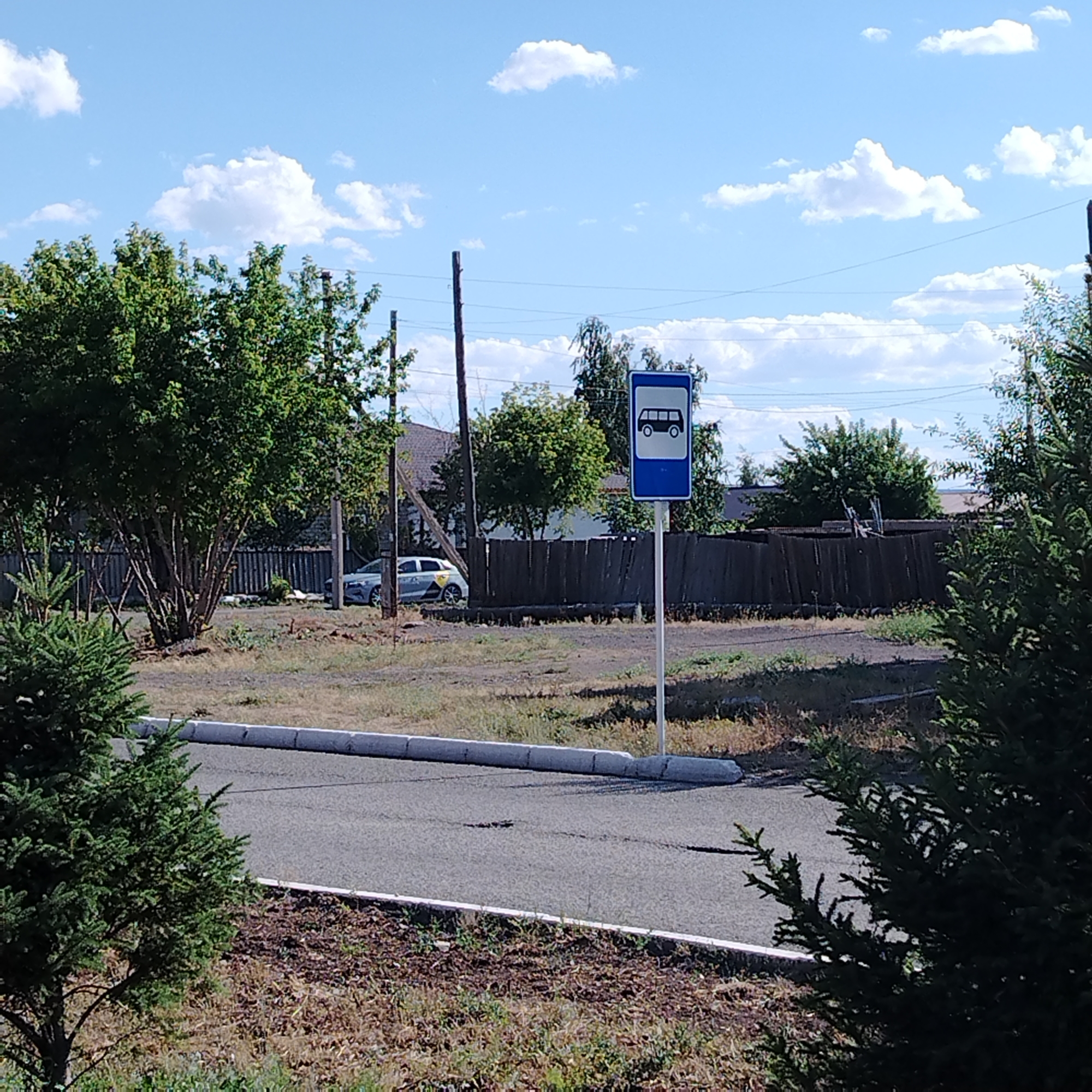 The image shows a bus stop sign on a street in the city of Ekibastuz, Kazakhstan. The sign is blue and white with a pictogram of a bus. A grey car is parked on the side of the road behind a wooden fence. There are trees and a few houses in the background. The sky is blue with white clouds. The ground is covered with dry grass and some weeds. The road in the foreground is paved and has a white curb.
