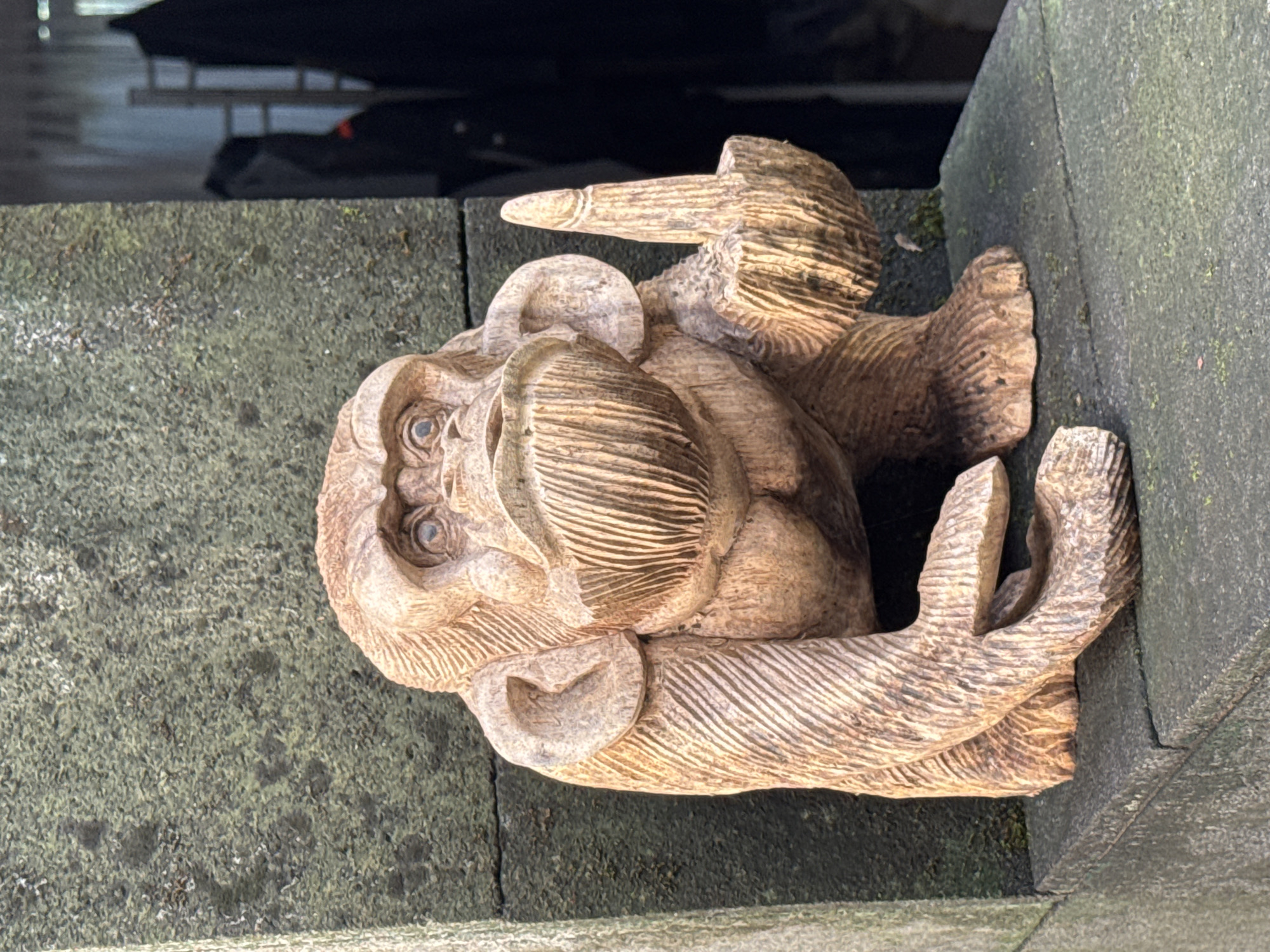A detailed, close-up shot depicts a carved wooden figure of a monkey placed on a mossy stone surface. The monkey figure, detailed with carved lines suggesting fur and facial features, including eyes, is positioned leaning against a grey stone wall. The image appears to have been taken outdoors, under natural light, capturing the textures of both the wooden carving and the stone surface. The background includes glimpses of dark, indeterminate objects and architectural elements. The subject is likely located in Ungasan, Indonesia.