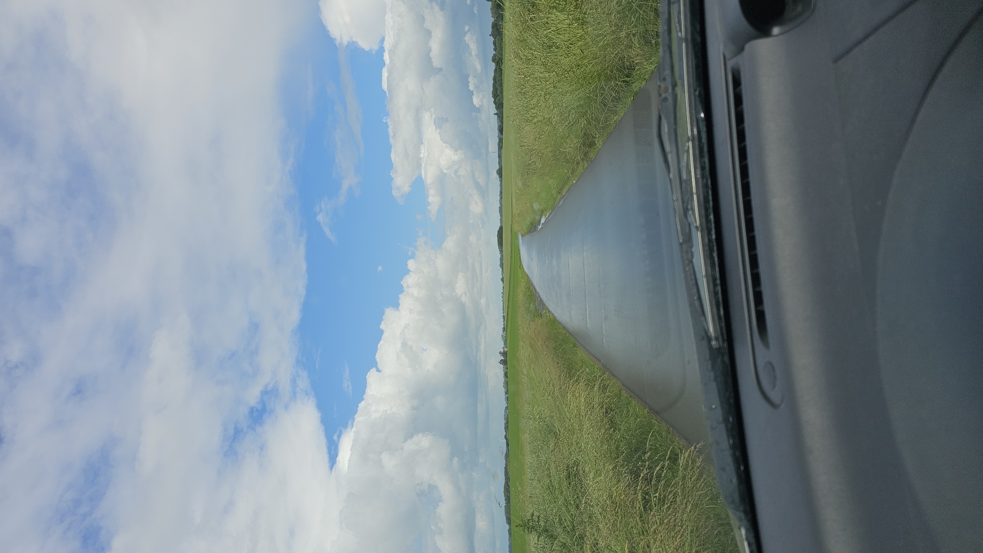 The image, taken from inside a vehicle looking forward, captures a serene daytime scene. A paved road or path stretches directly into the distance, appearing wet or reflective, and is bordered on both sides by lush, green grassy fields. Further in the background, a line of trees marks the horizon, suggesting a rural or countryside setting.
Above the landscape, a bright blue sky dominates, filled with large, fluffy white cumulus clouds. The illumination suggests a sunny day, likely mid-day, with good visibility.
On the right side of the frame, parts of the vehicle's dark gray dashboard and windshield wipers are visible, indicating the perspective is from the driver's or passenger's seat. There are no people visible, nor is there any discernible text within the image. The overall impression is one of calm and open space in a natural environment.