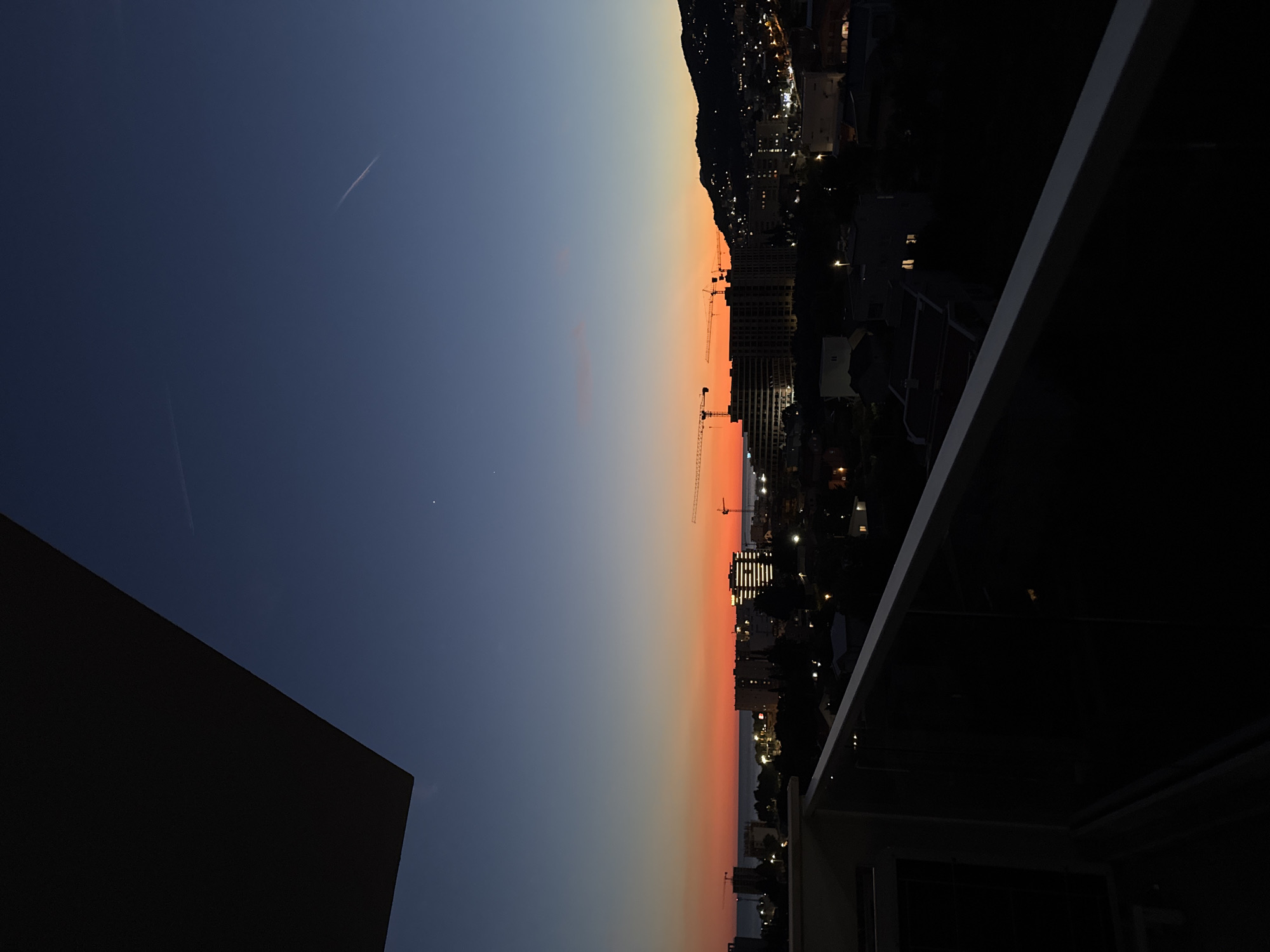 This is a medium shot, taken from a balcony, of a city skyline at dusk, likely in Bar, Montenegro. The sky transitions from a deep blue at the top to vibrant orange and red near the horizon. There are several construction cranes visible, suggesting ongoing development in the city. Buildings of various sizes are illuminated with lights, indicating an urban environment. The sea is visible in the distance, and a dark, mountainous silhouette forms the backdrop on the right side of the image. The foreground features the railing of the balcony, which is dark and partially obscures the view. No people or animals are visible in the frame.