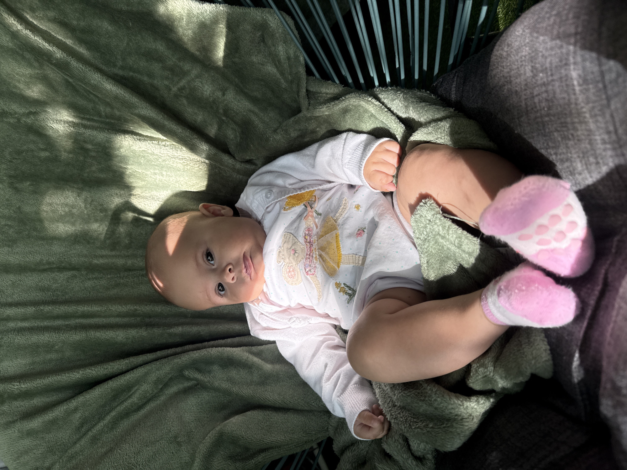 A baby is sitting on a green blanket, wrapped in a chair with blue stripes. The baby is wearing a white onesie with a drawing of a ballerina on it, and pink socks. The baby is looking at the camera with a serious expression. Sunlight is casting shadows on the baby and the blanket. There is no indication of location in Plovdiv, Bulgaria.
