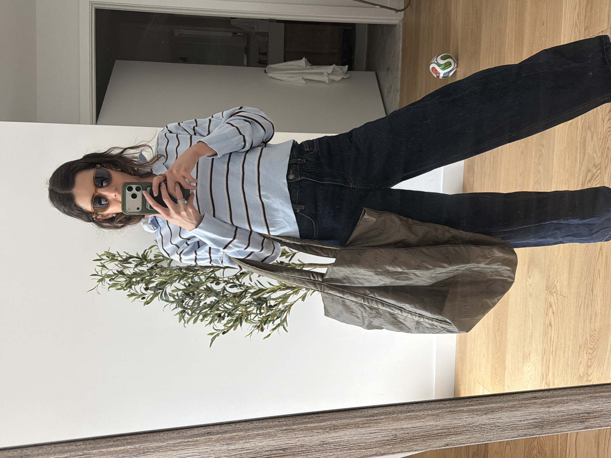 A full-body mirror selfie captures a young woman standing indoors. She has dark hair and wears brown sunglasses, with a smartphone in a green case held up to take the photo, obscuring her face. She is dressed in a light blue long-sleeved collared shirt with thin dark brown vertical stripes, paired with dark blue straight-leg jeans. Several rings adorn her fingers.
She carries a large, olive-green or dark brown tote bag on her right side. The bag features a subtle, repeated pattern across its surface, which appears to be a logo or text, though it is indistinct. A leafy green plant, resembling an olive branch, is positioned in the foreground to her left, partially obscuring the lower left portion of the image.
The setting is an interior space, likely a home or apartment, characterized by light wooden flooring and clean white walls. In the background, on the wooden floor to her right, a small, multi-colored ball is visible. A doorway is reflected behind her, showing a darker room beyond. The mirror itself has a rustic wooden frame at the bottom edge. The overall scene suggests a casual, everyday moment during daytime, potentially in New York, United States of America.