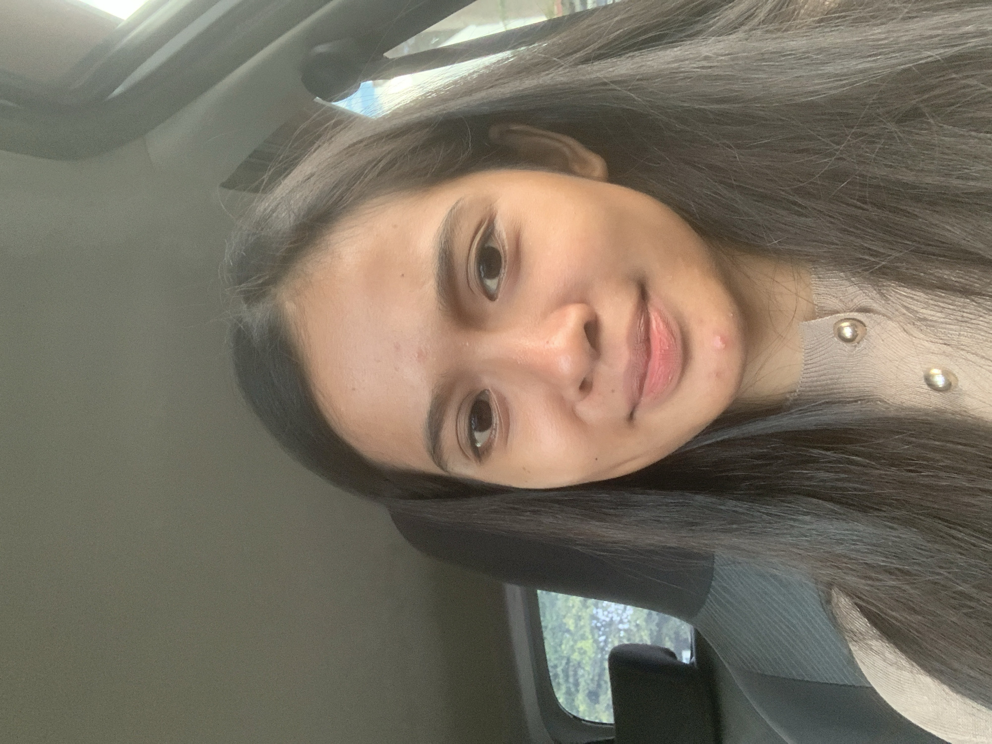 A woman with long, dark hair is seated in a car. The car's interior is visible, including part of the dashboard, a window with green foliage outside, and a sunroof. The woman is wearing a light-colored top with buttons. The location in Samarinda, Indonesia, is not determinable from the provided image.