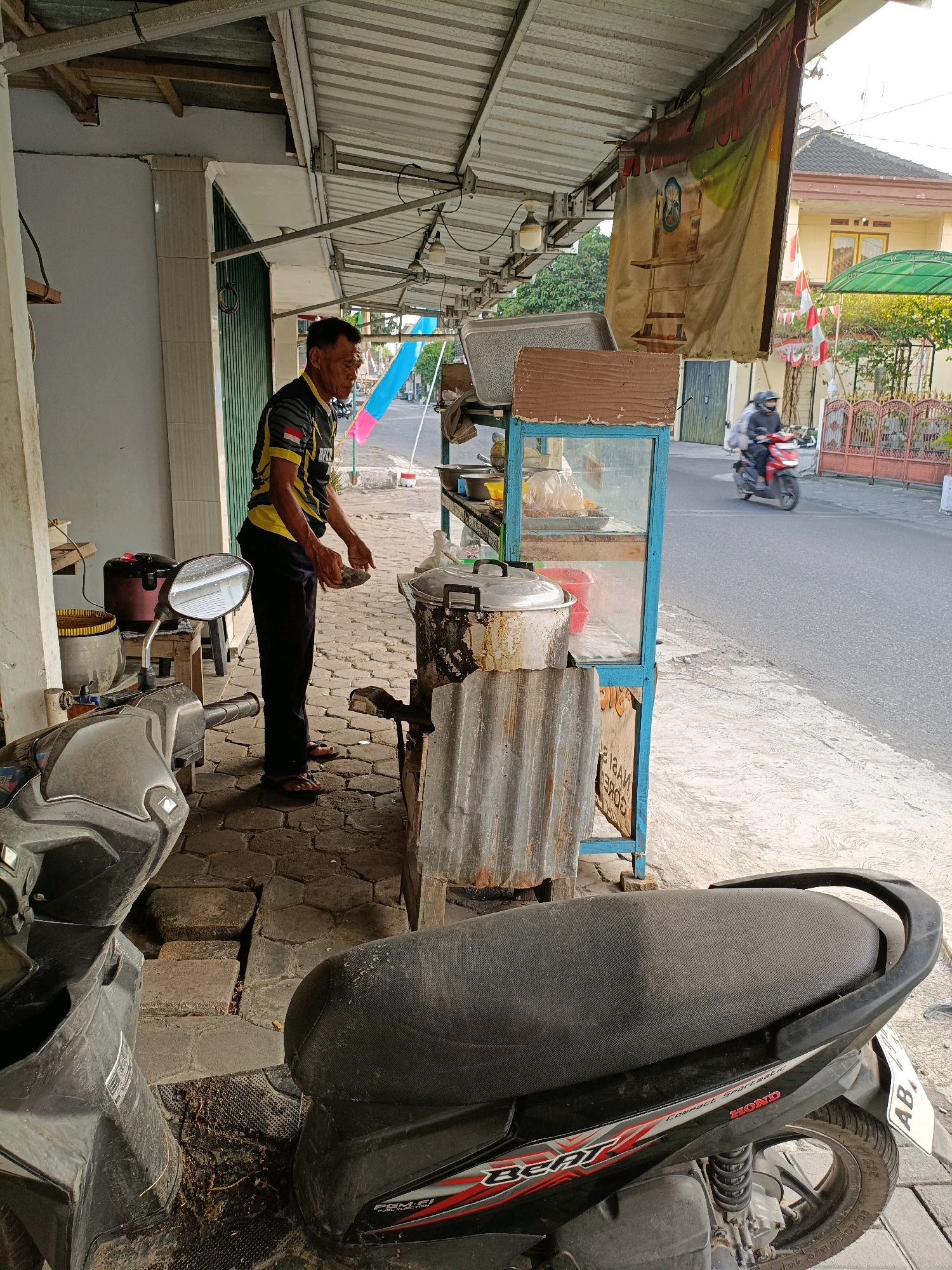 In Maguwoharjo, Indonesia, an outdoor scene captures a man standing by a food stall. He is wearing a black and yellow shirt with a white and red stripe on the arm and black pants. His hands are positioned as if he is holding or preparing food. Next to him is a food cart with a glass display showcasing food items. A large metal pot and other cooking utensils are visible. In the foreground, two dark-colored motorbikes are parked. The stall is situated on a paved walkway beside a road, with buildings and trees in the background. A person is riding a red motorbike on the road in the distance. The overall setting appears to be a casual street-side food vendor.