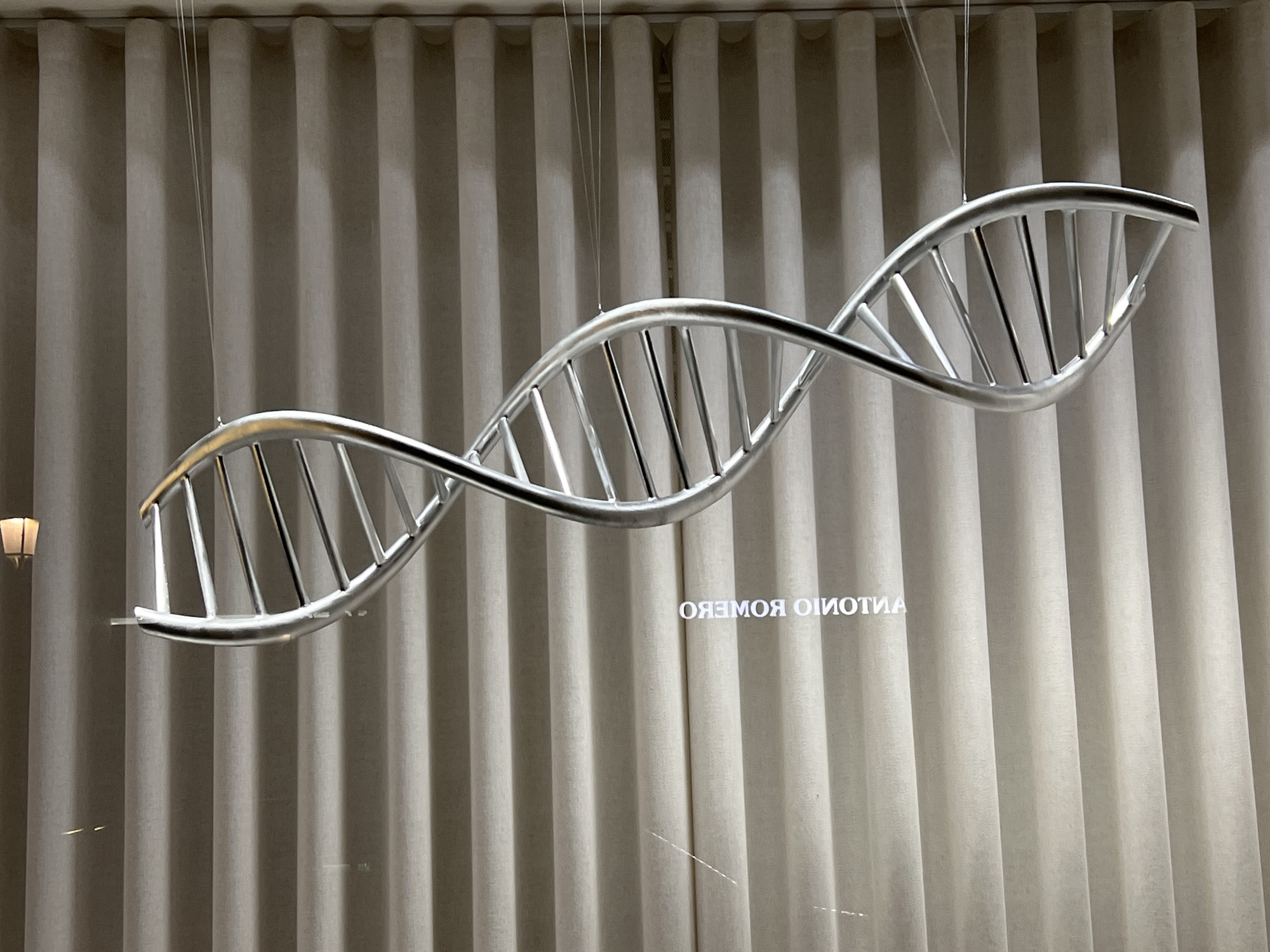 A silver DNA-shaped sculpture hangs in front of vertically draped light-colored curtains. Fine wires suspend the sculpture from the ceiling. Text, mirrored, is faintly visible through the curtains. The sculpture is likely located in València, Spain, based on the reverse spelling of "ANTONIO ROMERO." A lamp is partially visible in the left-hand corner of the image.