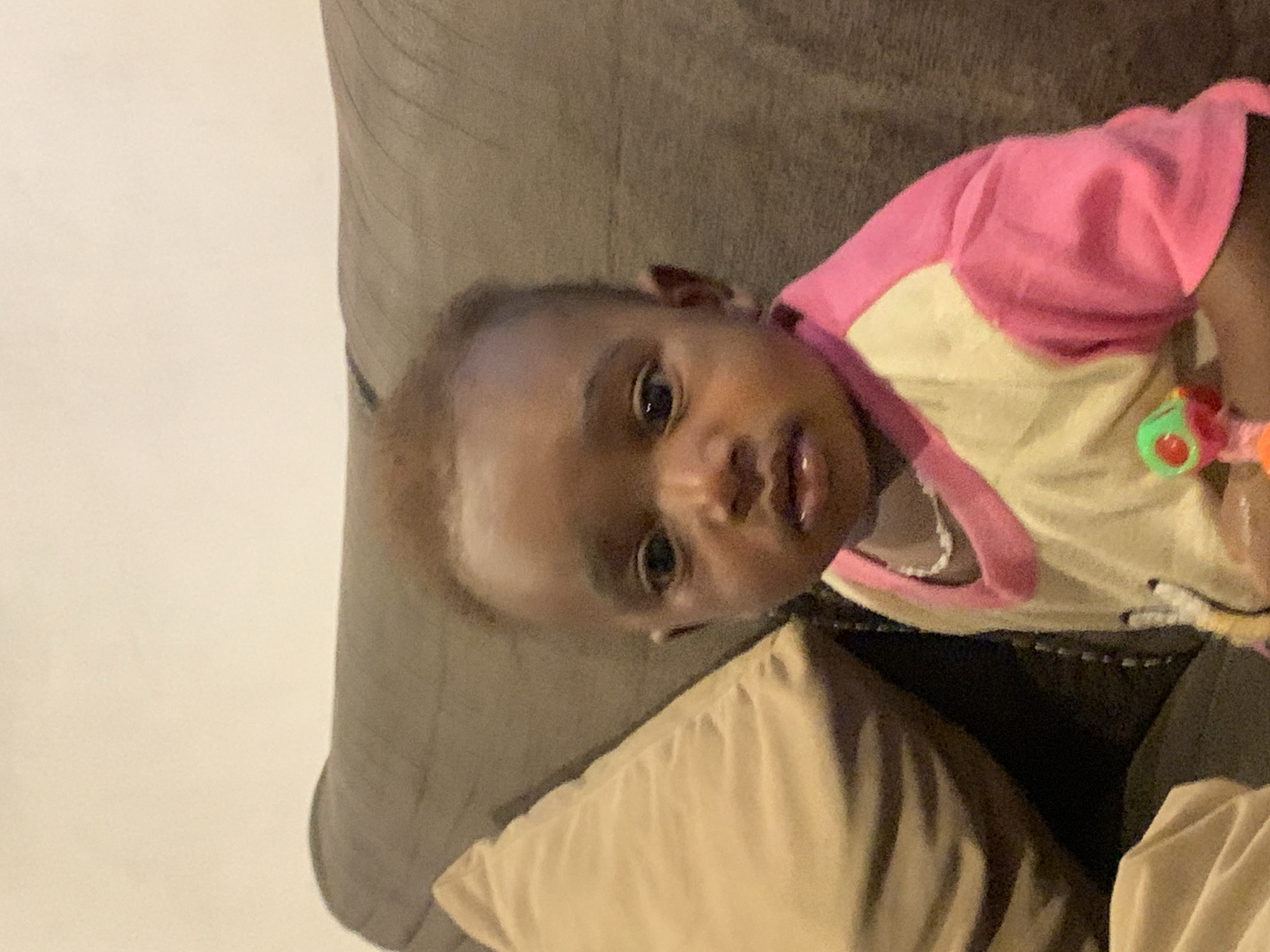 The image shows a baby with brown skin and short brown hair, lying on a tan pillow on a gray couch. The baby is wearing a pink and cream-colored shirt, with a toy in hand. The location in the city of Jos, Nigeria, cannot be determined from the image.