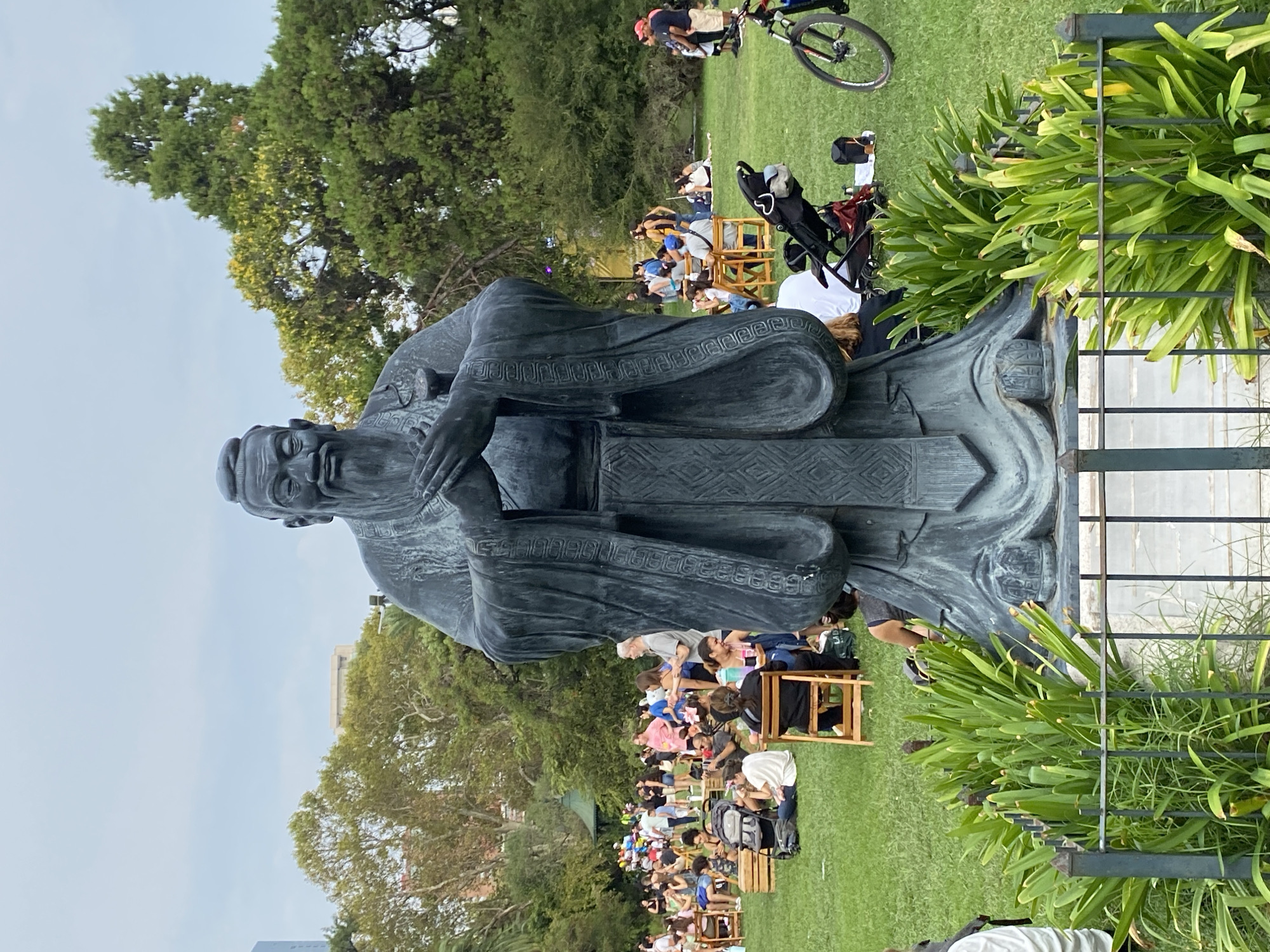A ground-level, eye-level, outdoor shot depicts a bronze statue of Confucius in a public park. The statue is the focal point of the image. The statue is a full-body sculpture of a man with long robes with detailed designs and facial features. The statue stands upright, with the figure's hands clasped in front of him. Behind the statue, there are many people of various ages sitting on chairs and on the grass, seemingly watching something. In the top right corner, a person on a bicycle is visible. To the right of the statue, plants and a metal fence are present. The backdrop includes trees and buildings. The scene appears to be located in Buenos Aires, Argentina, given the presence of the Confucius statue in Parque Lezama. The sky is partially visible in the background, suggesting an outdoor setting.