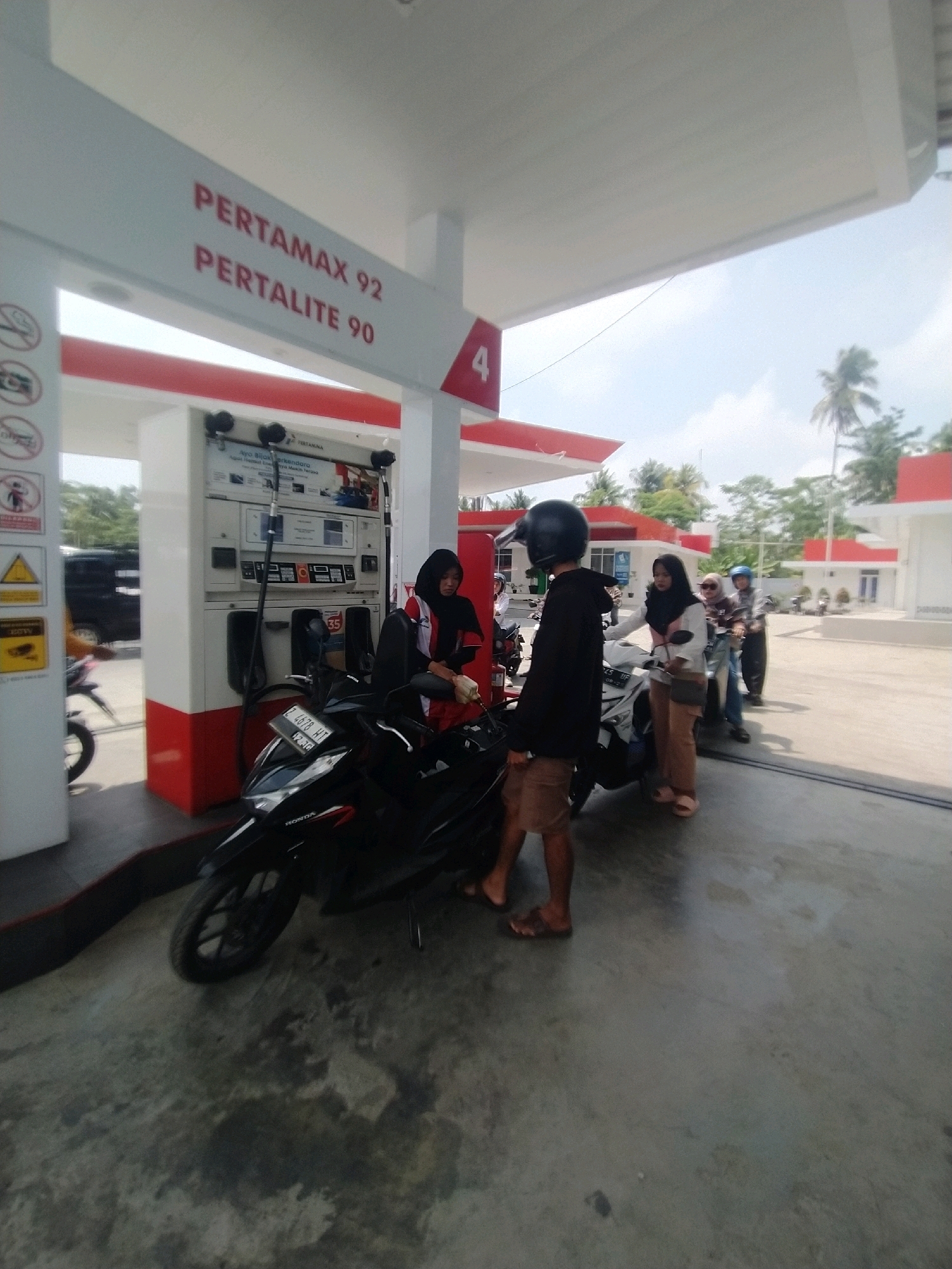 The image depicts a gas station in Desa Karangbenda, Indonesia, specifically a Pertamina station. Several people on motorcycles are present, appearing to be in the process of refueling or waiting. In the foreground, a black motorcycle is parked, and a person wearing a black shirt and shorts stands beside it. Further back, other individuals are seen on their motorcycles, including a woman in a black hijab attending to a pump. The gas station has a white canopy with red accents, and signage indicates "PERTAMAX 92" and "PERTALITE 90." The environment is outdoors, with a bright, sunny sky and some palm trees and buildings visible in the background, suggesting a tropical or sub-tropical climate. The ground is paved concrete.