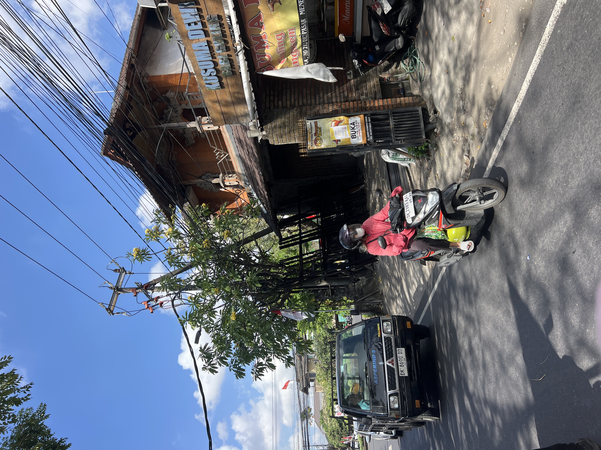 This image captures a street scene in Denpasar, Indonesia, likely on a sunny day. A black utility truck with the license plate "DK 8690 DD" is prominent in the foreground, traveling on the right side of the road. Beside it, a person on a red scooter, wearing a helmet and a pink jacket, is also moving forward. Behind the scooter, a black motorcycle is parked.
The background reveals a traditional Indonesian building with an orange facade and a terracotta roof, partially obscured by a large tree with yellow flowers. A sign with the text "KUSUMA DEWI BABI GULING" is visible on the building, indicating it's a restaurant serving roasted suckling pig. Another sign, "BUKA" (Open) with operating hours and address, is displayed near the entrance.
Electrical wires are strung across the scene, supported by utility poles. Two flags, one red and white (likely the Indonesian flag) and another white flag, are seen fluttering on poles in the distance. The road is paved with asphalt and has a white lane marker. Shadows of trees and vehicles are cast on the road, indicating the time of day. The environment appears to be urban or suburban, with a mix of commercial establishments and residential buildings.