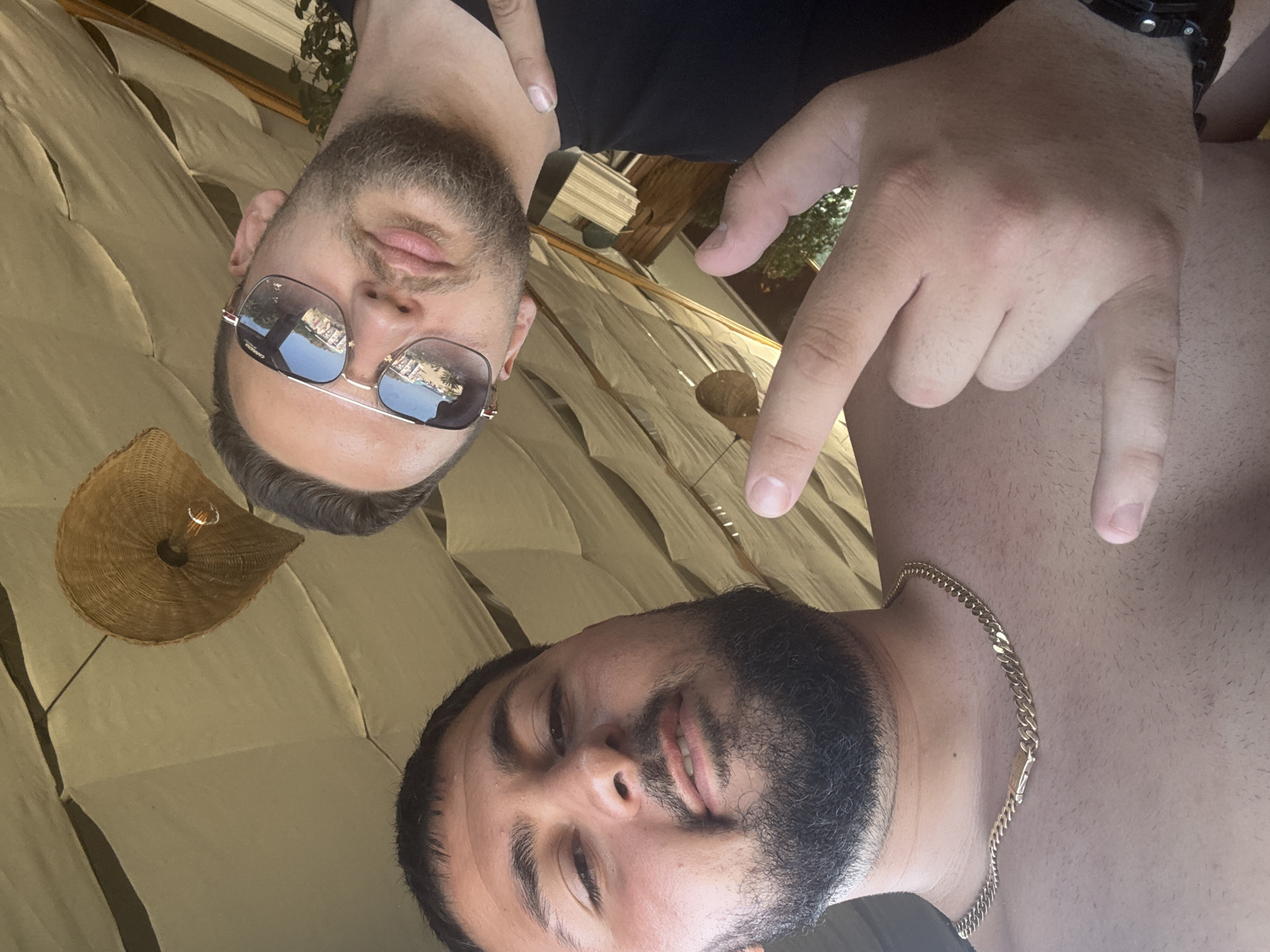 Two men are captured in a close-up selfie. The man in the foreground, on the right, is shirtless and wears a gold chain. His head is tilted, and he is looking directly at the camera with a slight smile. The man in the background, on the left, is wearing sunglasses and a black shirt. He is also looking at the camera. His lips are slightly parted, and he has a beard and stubble. A wicker lampshade with a lightbulb is visible in the background. The overall impression is casual and relaxed, possibly taken on a sunny day. The image appears to be taken outdoors, given the lighting and the background elements. The location context provided is Sinemorets, Bulgaria, which could suggest a vacation or a day out at the beach or a resort. The lighting suggests it is daytime.