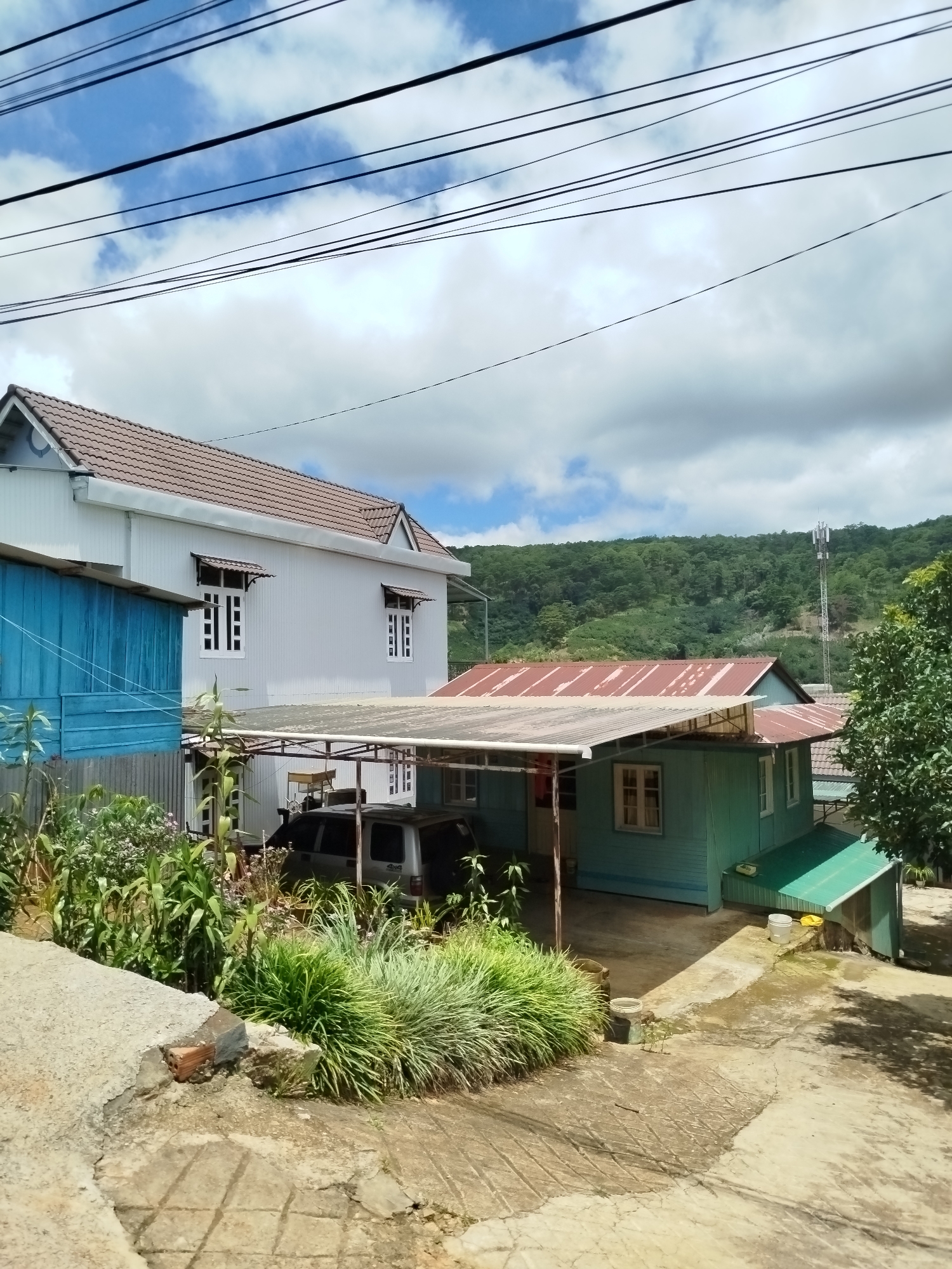 The image depicts a rural residential area in Xuân Sơn, Vietnam, on a cloudy day. In the foreground, a cracked concrete pathway leads to a yard with lush greenery. Two houses are visible: a larger, white, two-story house with a brown tiled roof on the left, and a smaller, teal-colored house with a red corrugated metal roof on the right. A blue structure is partially visible to the left of the white house. A white minivan is parked under a carport attached to the teal house. In the background, rolling green hills covered in trees rise under a sky with scattered clouds, and a cell tower stands prominently on one of the hills. Overhead, multiple power lines crisscross the frame.