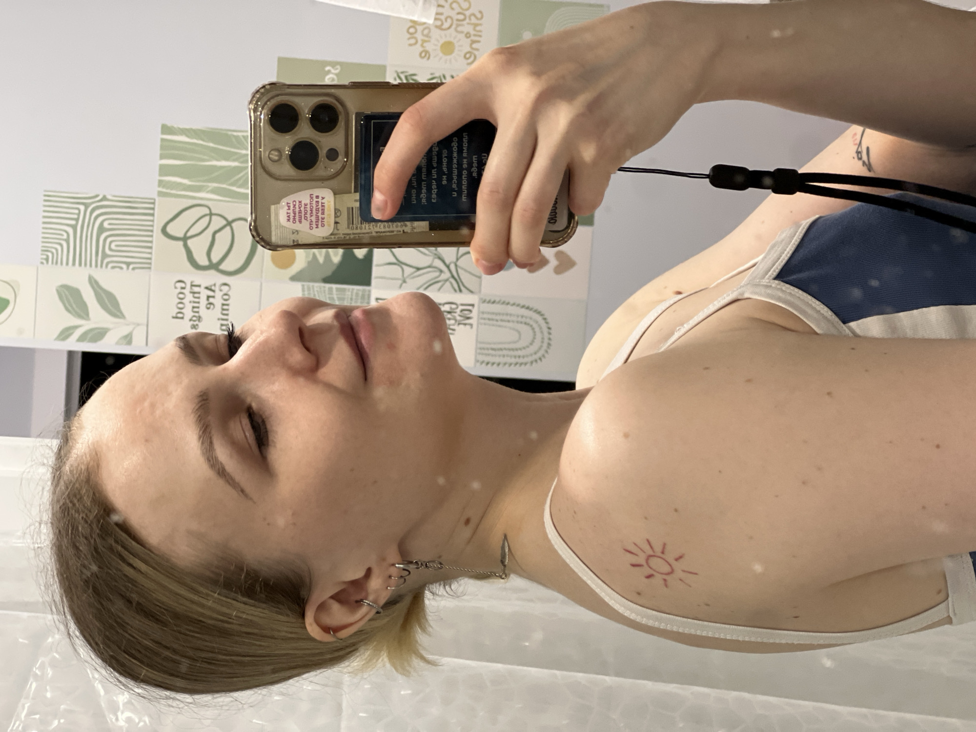 A pale-skinned woman is holding a smartphone to take a selfie. She is wearing a white tank top with blue trim and has a small sun tattoo on her right shoulder. She has multiple piercings in her left ear and a thin silver chain connecting two of them. The phone case is clear with gold accents and contains what appears to be a Russian book insert with text in Cyrillic script. Part of the text on the book insert visible in the frame includes phrases like "чтоте ОТР УМОТОп," "оте RAst Y," and "ил онл". The background consists of white walls with a strip of decorative tiles with light green patterns along the top. The OCR text on the phone case indicates the location Khimki, Russia.