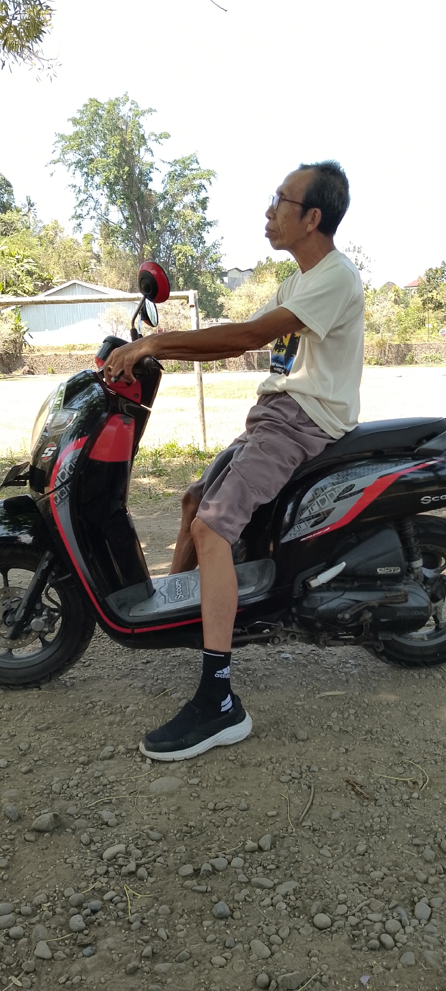 An East Asian man, wearing glasses, a light-colored t-shirt, gray shorts, black socks with white stripes, and black slip-on shoes, is seated on a black and red scooter in an outdoor, rural setting. The scooter has "SCOOPY" written on the footrest. The man's left hand rests on the handlebar. The background features trees, vegetation, and buildings, possibly indicating a location in Pekutatan, Indonesia. The ground is covered with gravel and dirt. The lighting suggests a daytime scene.