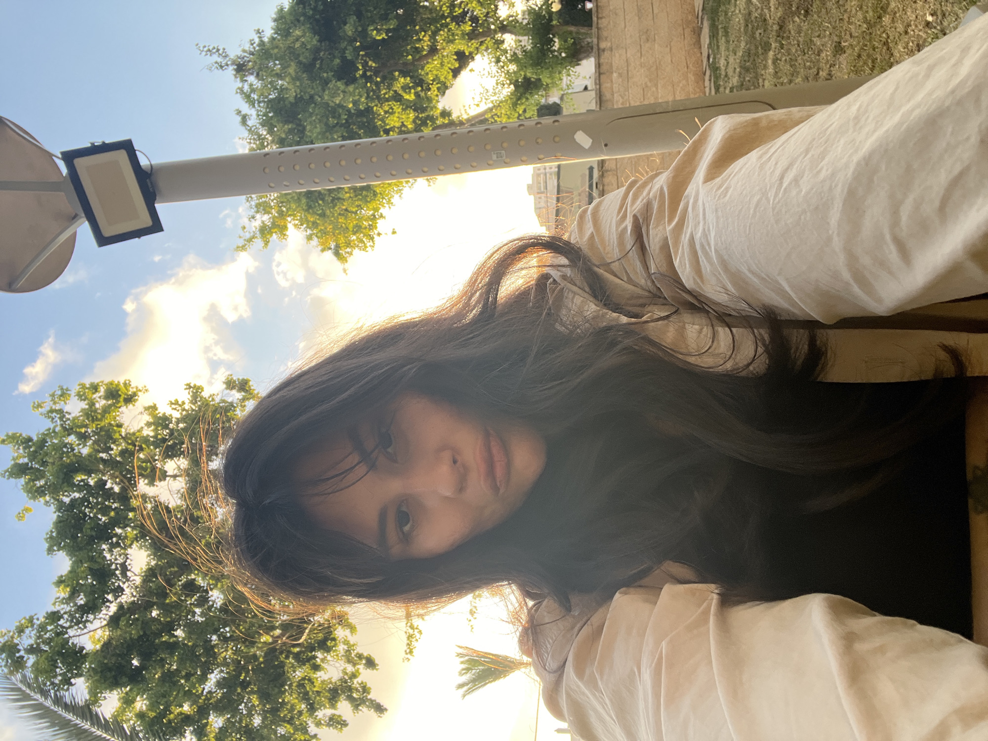 A young woman is taking a selfie outdoors in Kiryat Bialik, Israel. She has long, dark hair that frames her face and is wearing a light-colored shirt. The sun is shining brightly, casting a warm glow on her and the scene. Behind her, lush green trees and a bright blue sky with scattered clouds are visible. A white pole with a light fixture is also present in the frame. The overall mood appears relaxed and pleasant, suggesting a casual outing on a sunny day. The image captures a moment of personal reflection or a casual share with friends.