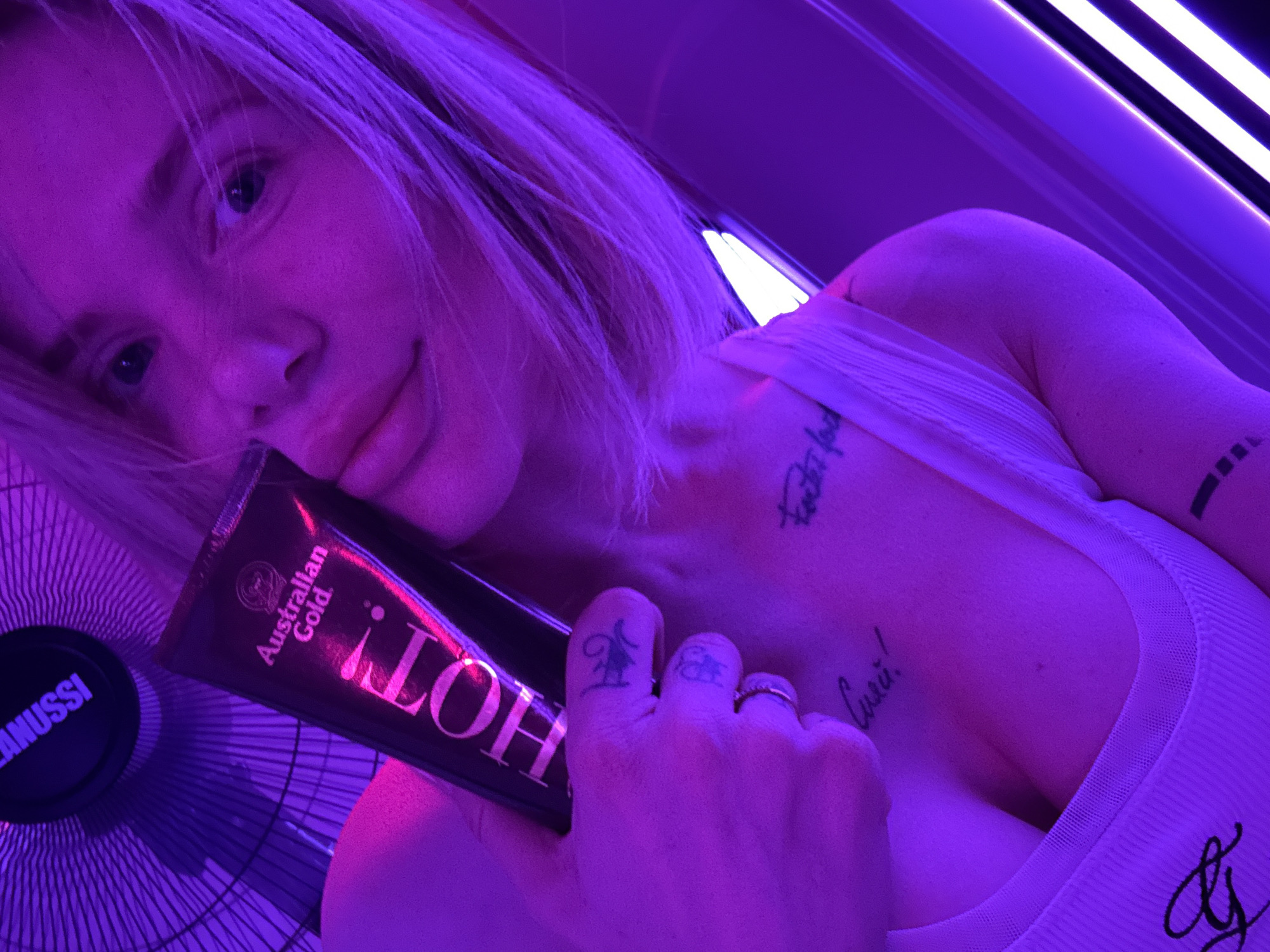 A blonde woman with light skin poses in a tanning bed. She is holding a tube of Australian Gold "HOT!" tanning lotion. Her hand, adorned with rings and tattoos, is visible. She has tattoos on her chest and arm. A Janussi fan is in the background. The environment is lit with purple and white light. There is no indication in the image of the location as Gorno-Altaysk, Russia.