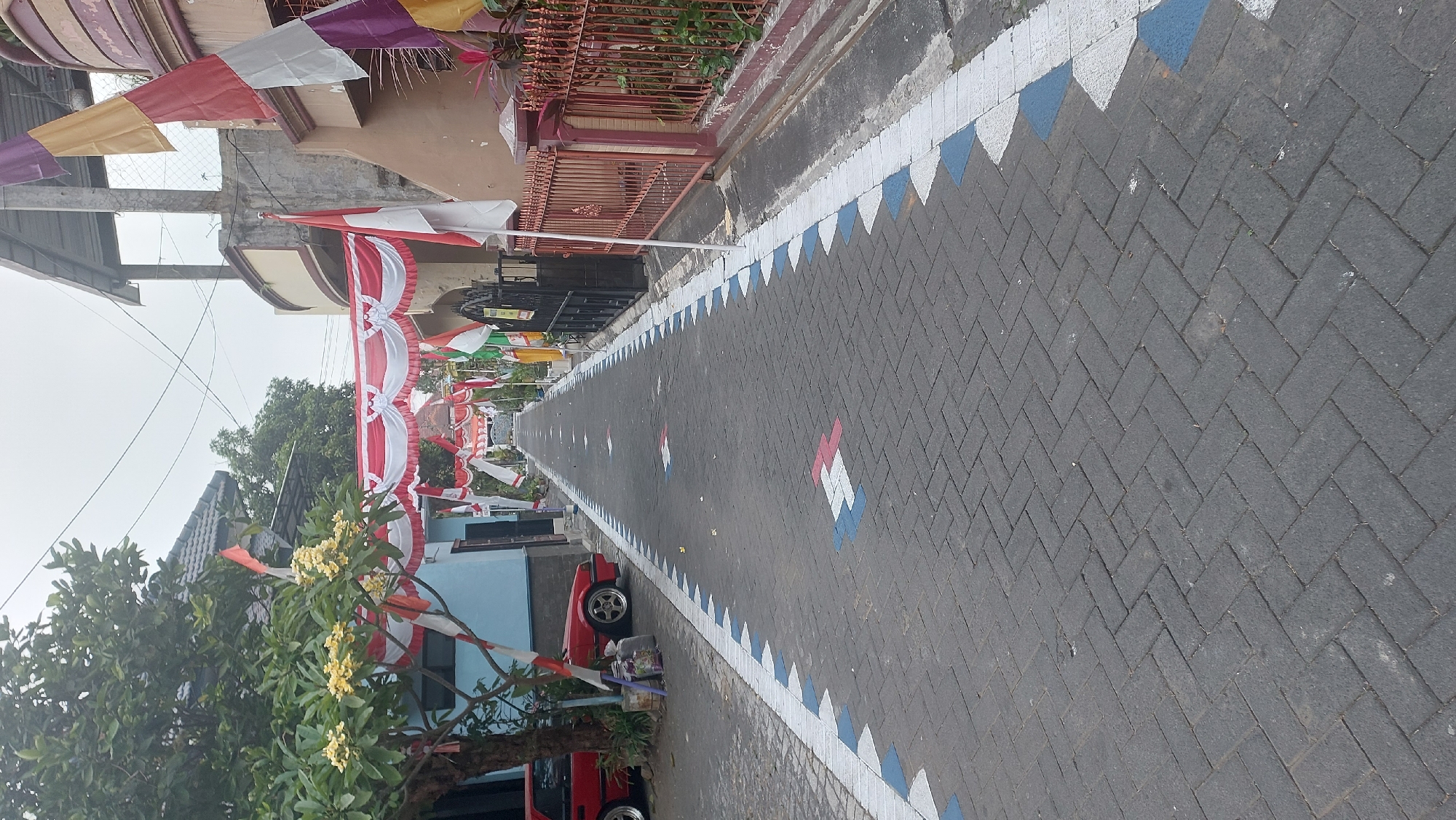 The image captures a street in Malang, Indonesia, decorated for a national celebration, likely Independence Day. The street is paved with interlocking bricks and is lined with red and white banners and flags, including the Indonesian flag. Along the sides of the street, there are houses with fences. Some of the brick paving has been painted with decorative patterns in red, white, and blue, forming a border along the edge and repeating patterns on the road itself. A red car is parked on the left side of the street, near a tree with yellow flowers. The sky is overcast.