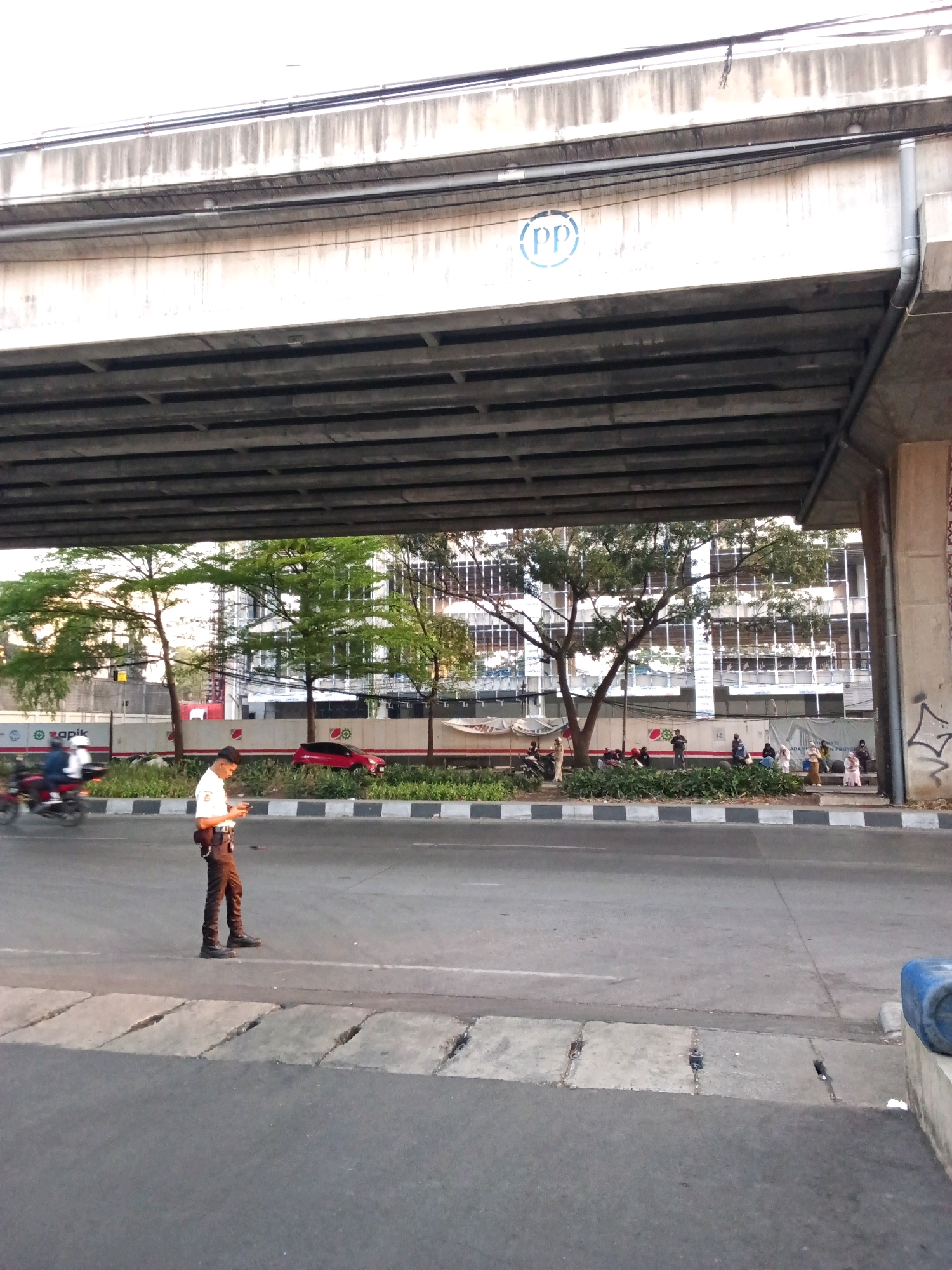The image depicts a street scene under an elevated structure, likely an overpass or bridge. A lone individual, dressed in a white shirt and dark pants, stands on the asphalt road, looking at a mobile phone. The person appears to be a traffic controller or security personnel due to the attire and the location. Several trees with green foliage are visible on the side of the road, with a building in the background featuring a modern architectural style with many windows. A red car is parked near the trees, and a motorcycle with two occupants is in motion on the left side of the frame. The elevated structure has a symbol of "(PP)" stenciled in blue on its side. The environment suggests an urban setting with ongoing traffic and pedestrian activity. The image appears to be taken in Bandung City, Indonesia, given the context provided.